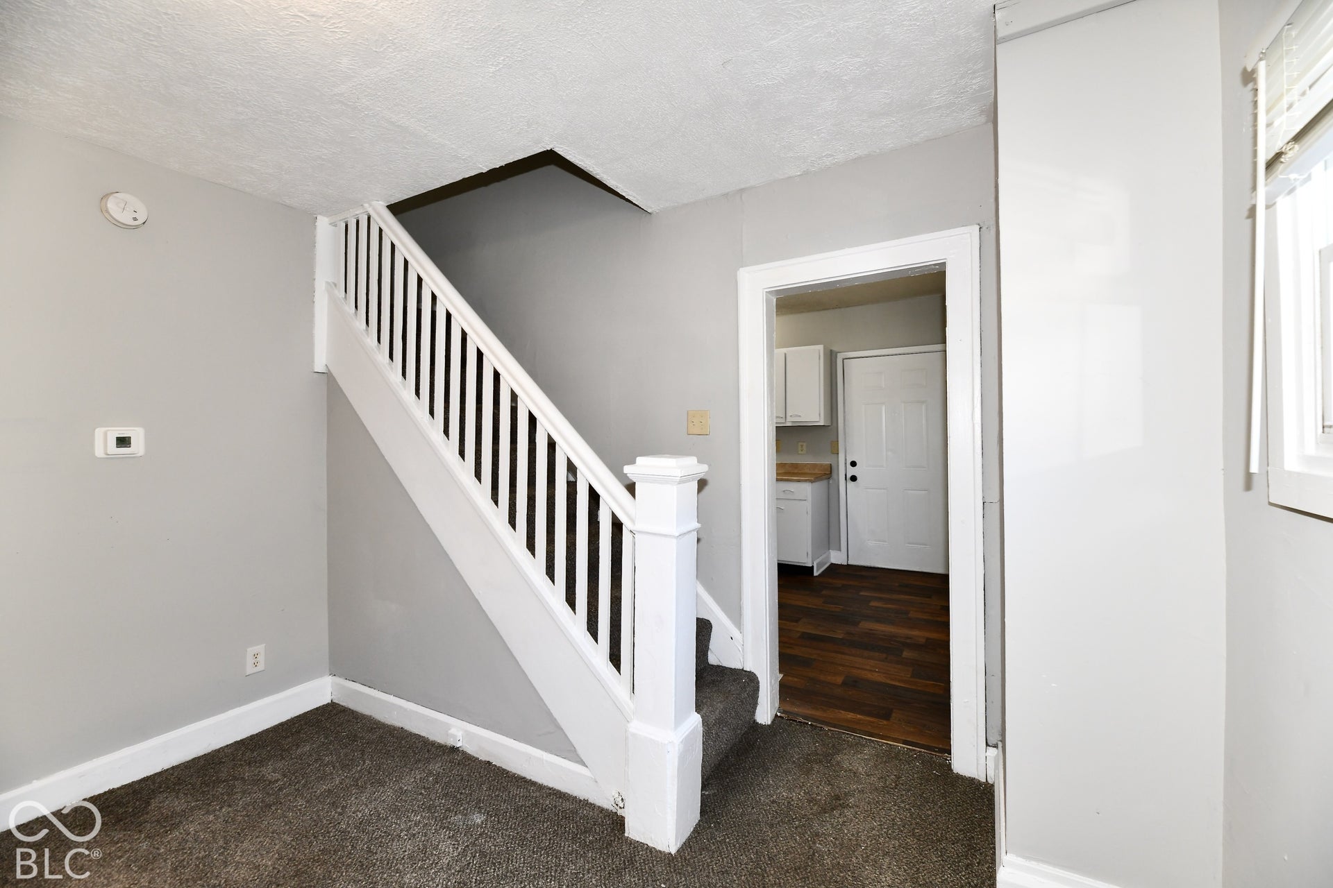 Carpeted flooring, white painted staircase with balusters, light gray wall paint, doorway with white trim, wood-finish flooring visible through doorway