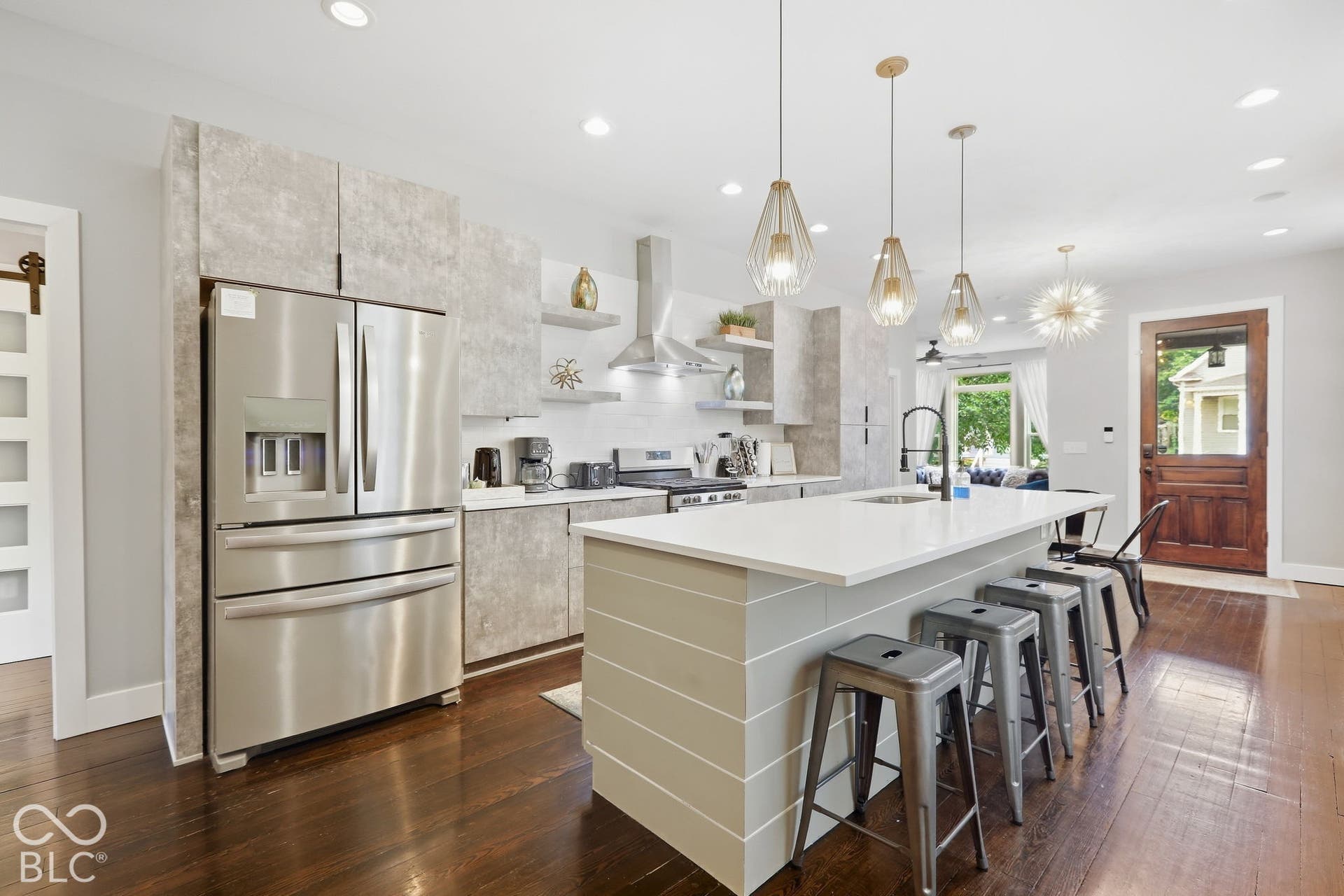 Stainless steel French door refrigerator, white kitchen island with shiplap detailing, dark wood-finish flooring, three pendant lights, and a white tiled backsplash