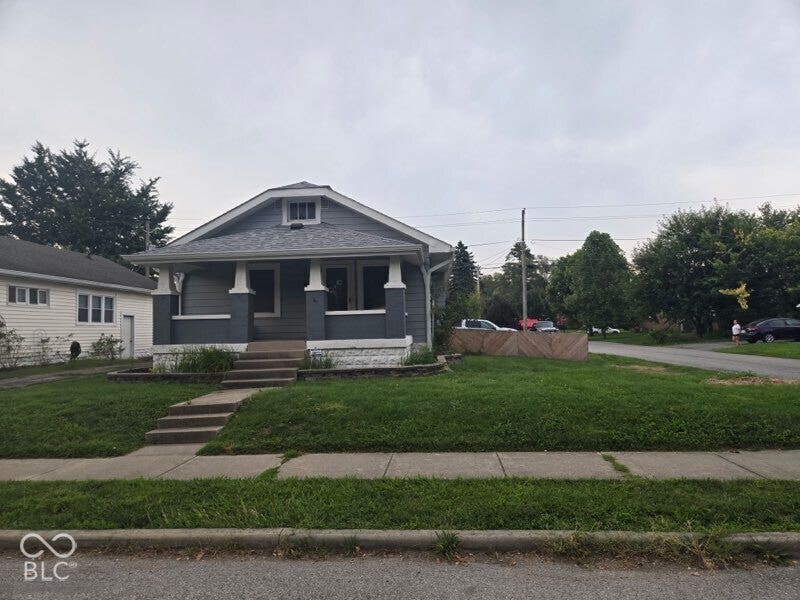 Charming brick-and-siding home featuring a distinctive dark exterior with gabled roofline, front-facing covered porch with pendant lighting, concrete foundation, and manicured front lawn with mature landscaping and established trees