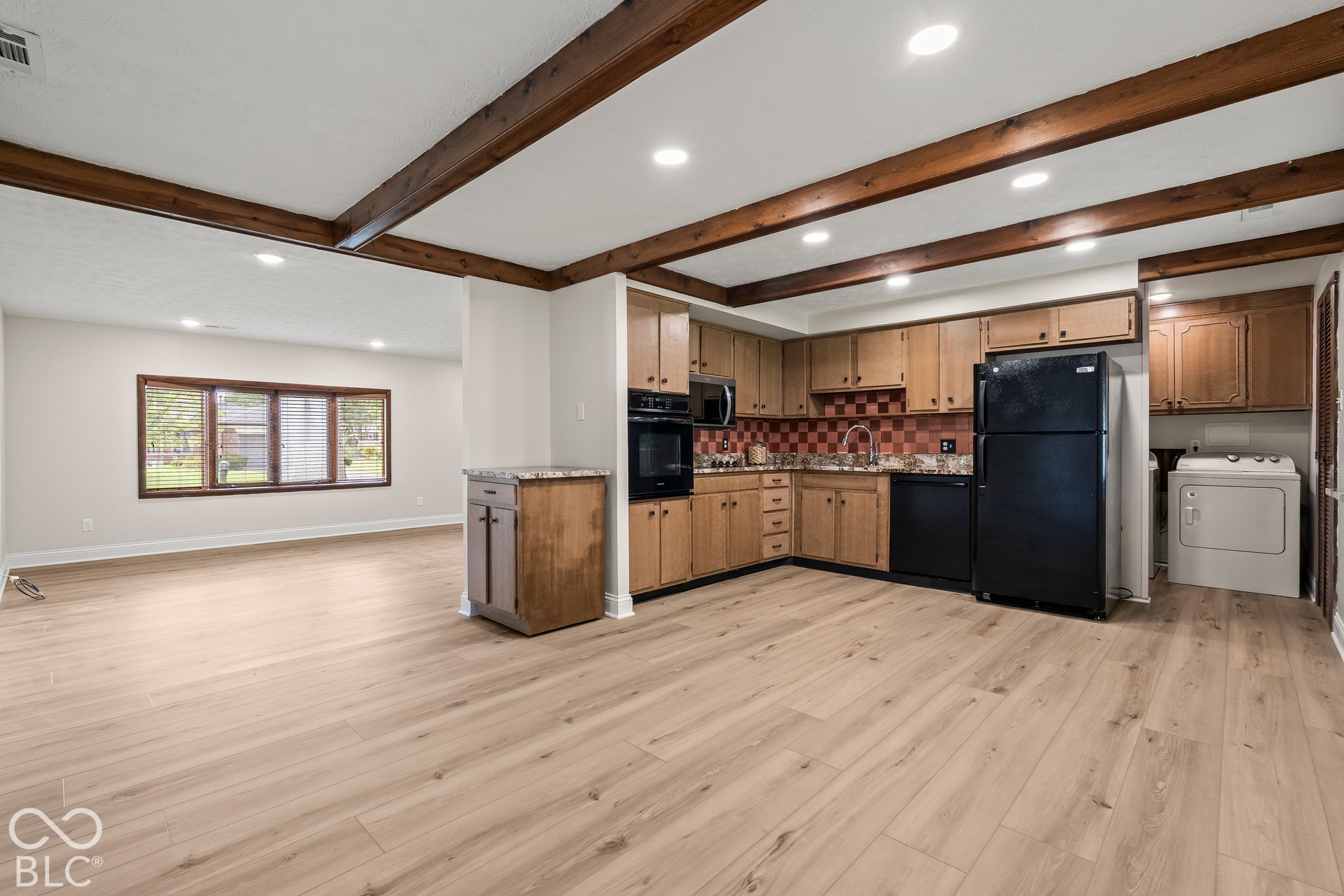 Spacious open-concept area featuring wood-finish flooring, exposed wood ceiling beams, recessed lighting, and a kitchen with wood cabinetry
