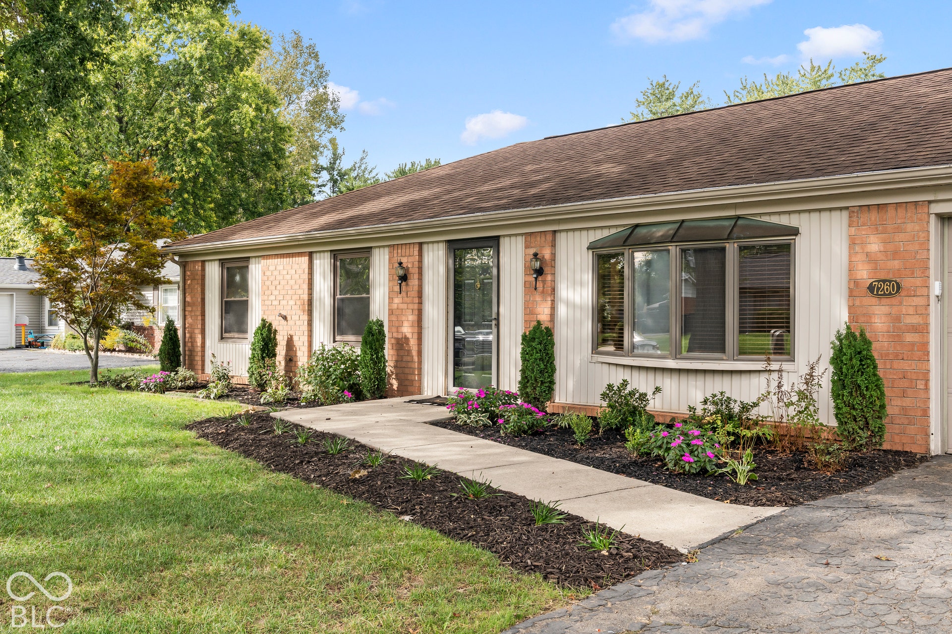 Exterior featuring brick and vertical siding, concrete walkway, bay window with a protective awning, and mature landscaping
