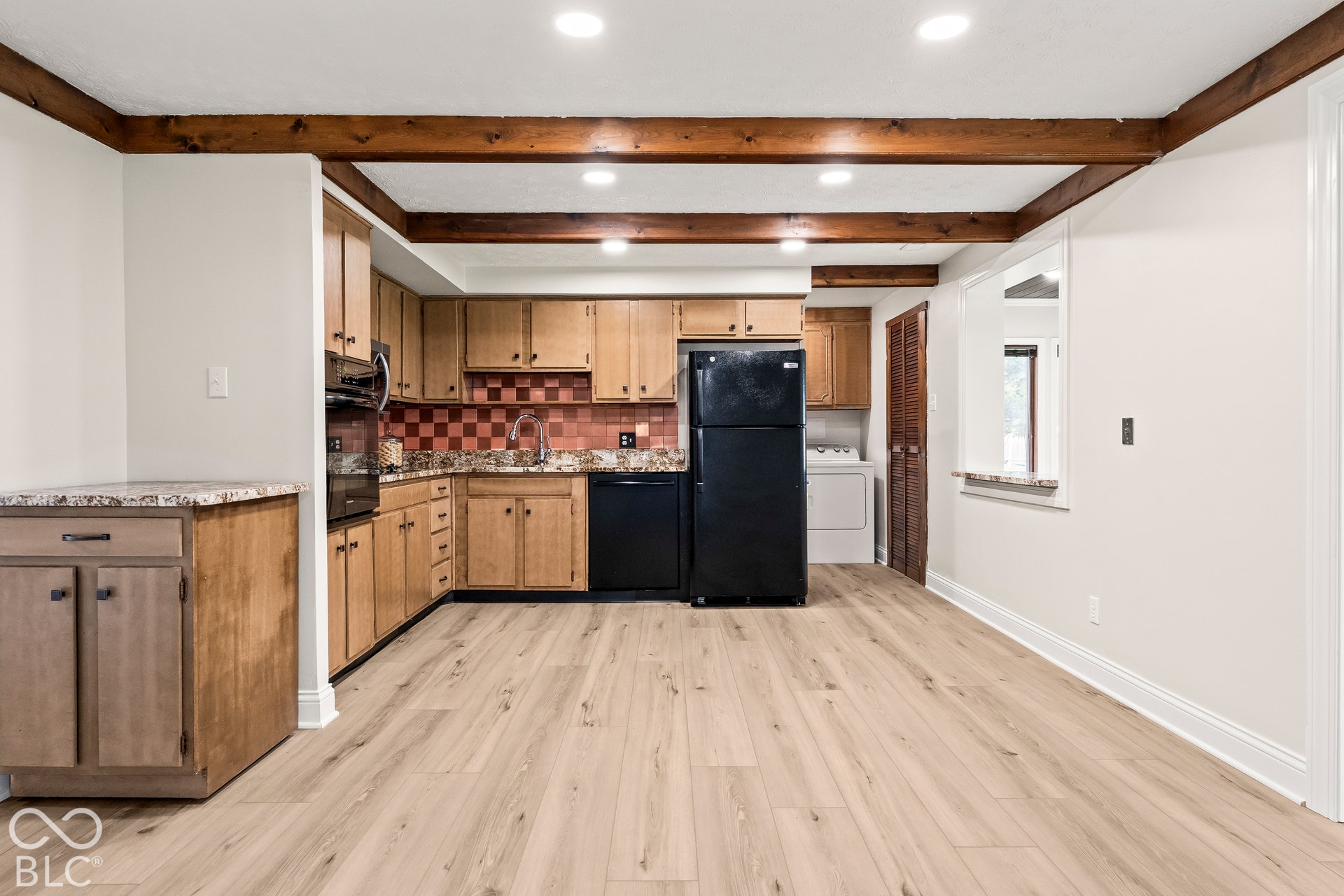 Wood-finish flooring, exposed ceiling beams, light wood cabinetry, checkerboard tile backsplash, and a pass-through opening