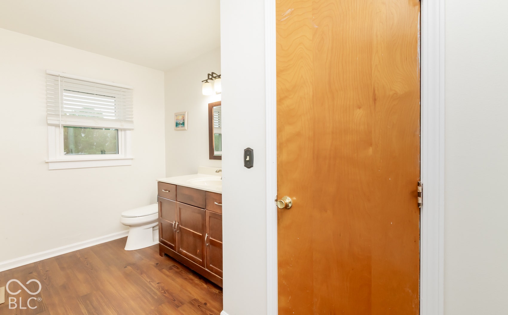 Wood-finish flooring with a white toilet, dark wood-tone vanity, and a window featuring blinds