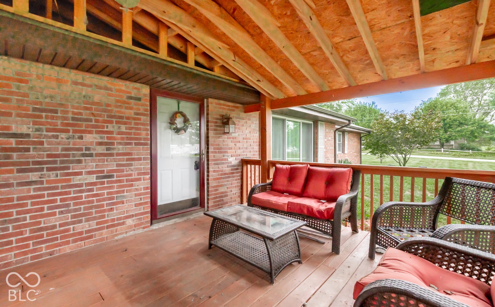 Covered wood-finish porch featuring a red brick facade, exposed wood ceiling beams, wood-finish railing, and an exterior light fixture