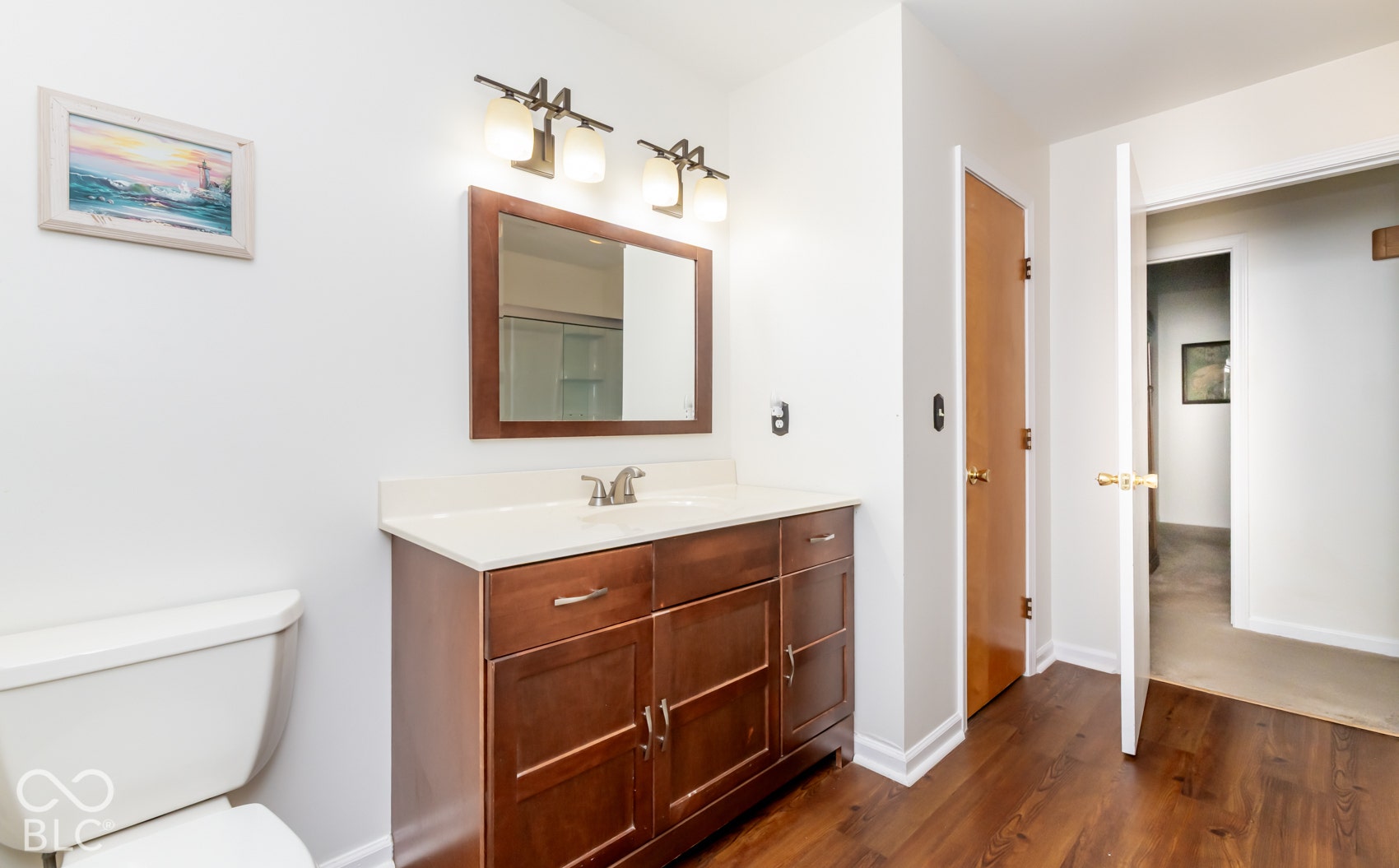 Spa-inspired primary bathroom featuring a wood-finish vanity with dual drawers and chrome hardware, white quartz countertop, polished chrome faucet and fixture, framed mirror with integrated lighting, and warm wood-finish flooring throughout