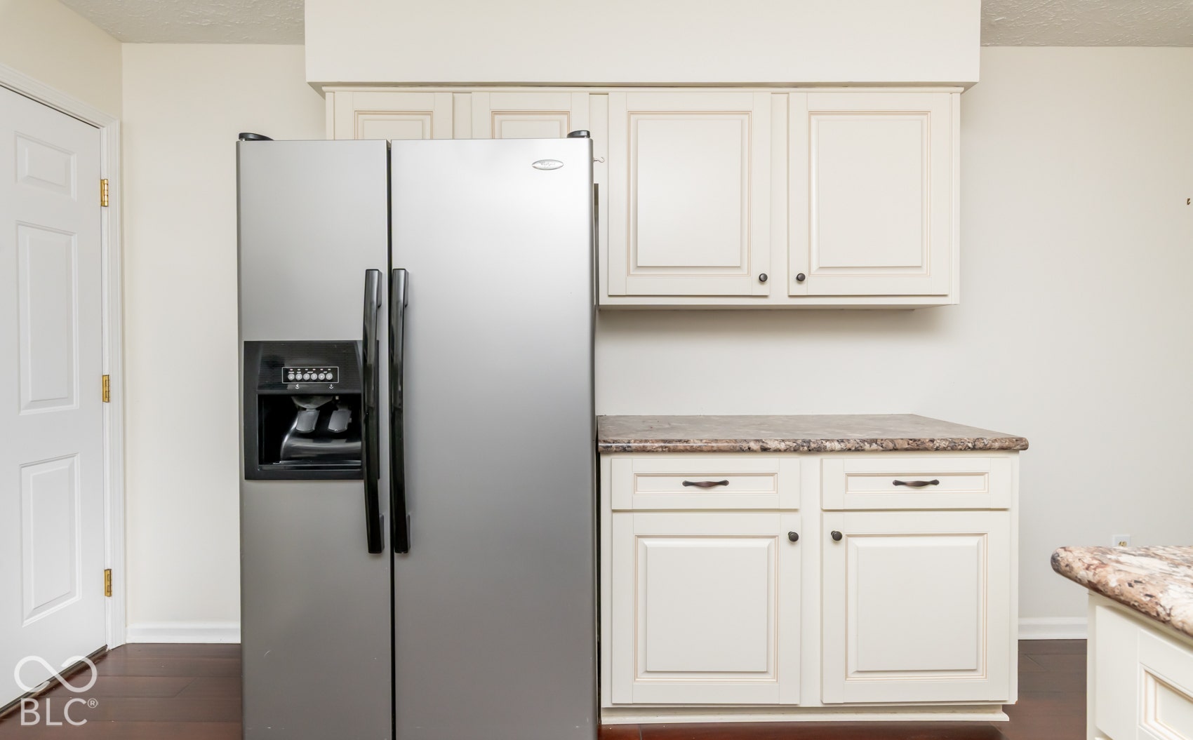 Stainless steel side-by-side refrigerator, cream cabinetry, dark wood-finish flooring, and light-toned countertops