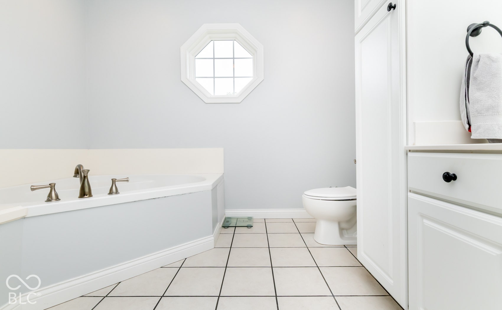 Soaking tub with bronze-finish fixtures, octagonal window, white tile flooring, built-in white cabinetry, and white counter surfaces