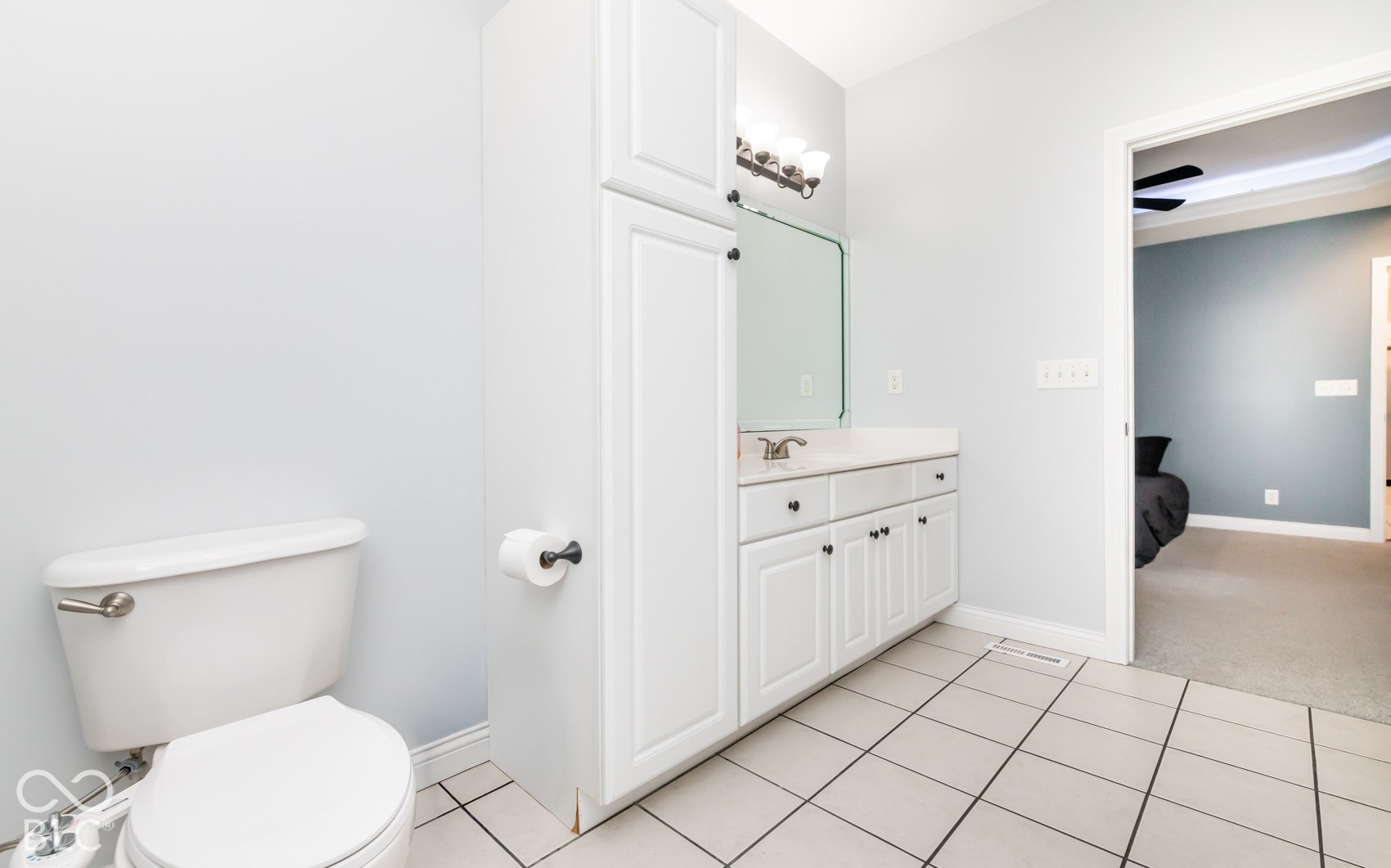 White tile flooring, white vanity with integrated sink, rectangular mirror, light fixture with multiple bulbs, and white built-in storage cabinetry