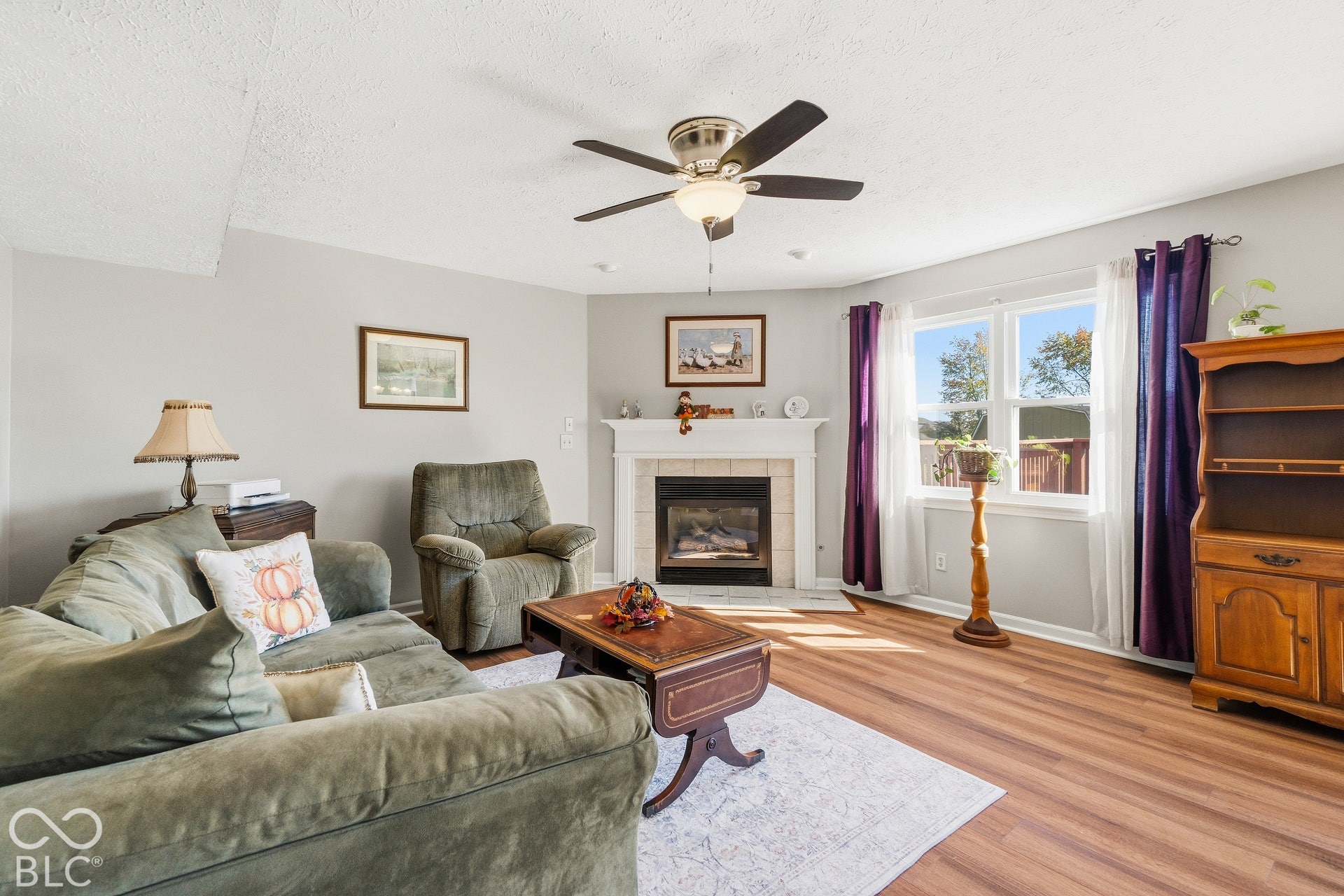 Living area featuring wood-finish flooring, a white-mantled fireplace, and a ceiling fan with integrated lighting