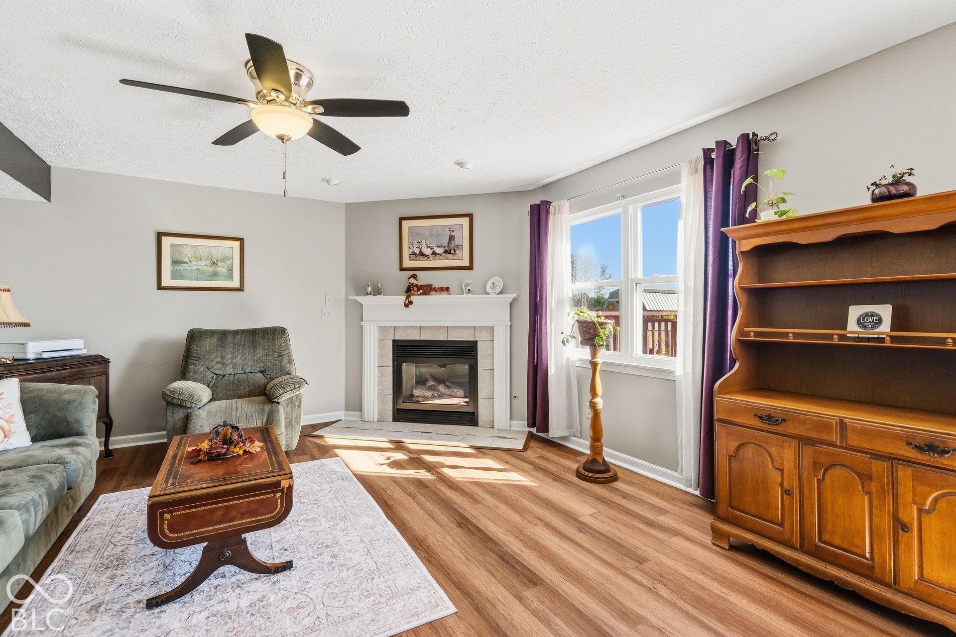 Wood-finish flooring, white fireplace with tile surround, white window trim, and a ceiling fan with light fixture