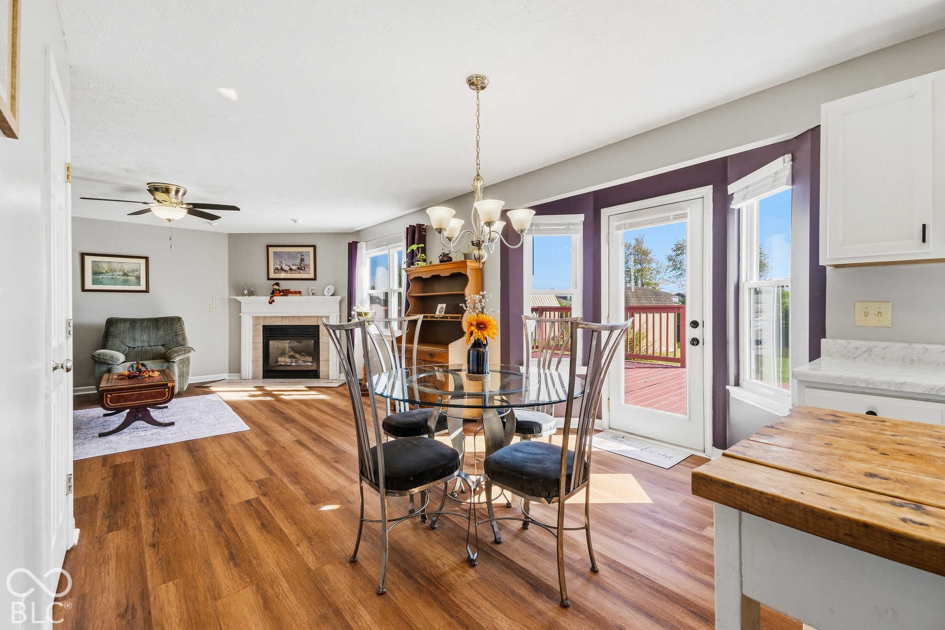 Wood-finish flooring throughout, with a fireplace and white mantel, a ceiling fan, and a chandelier lighting fixture