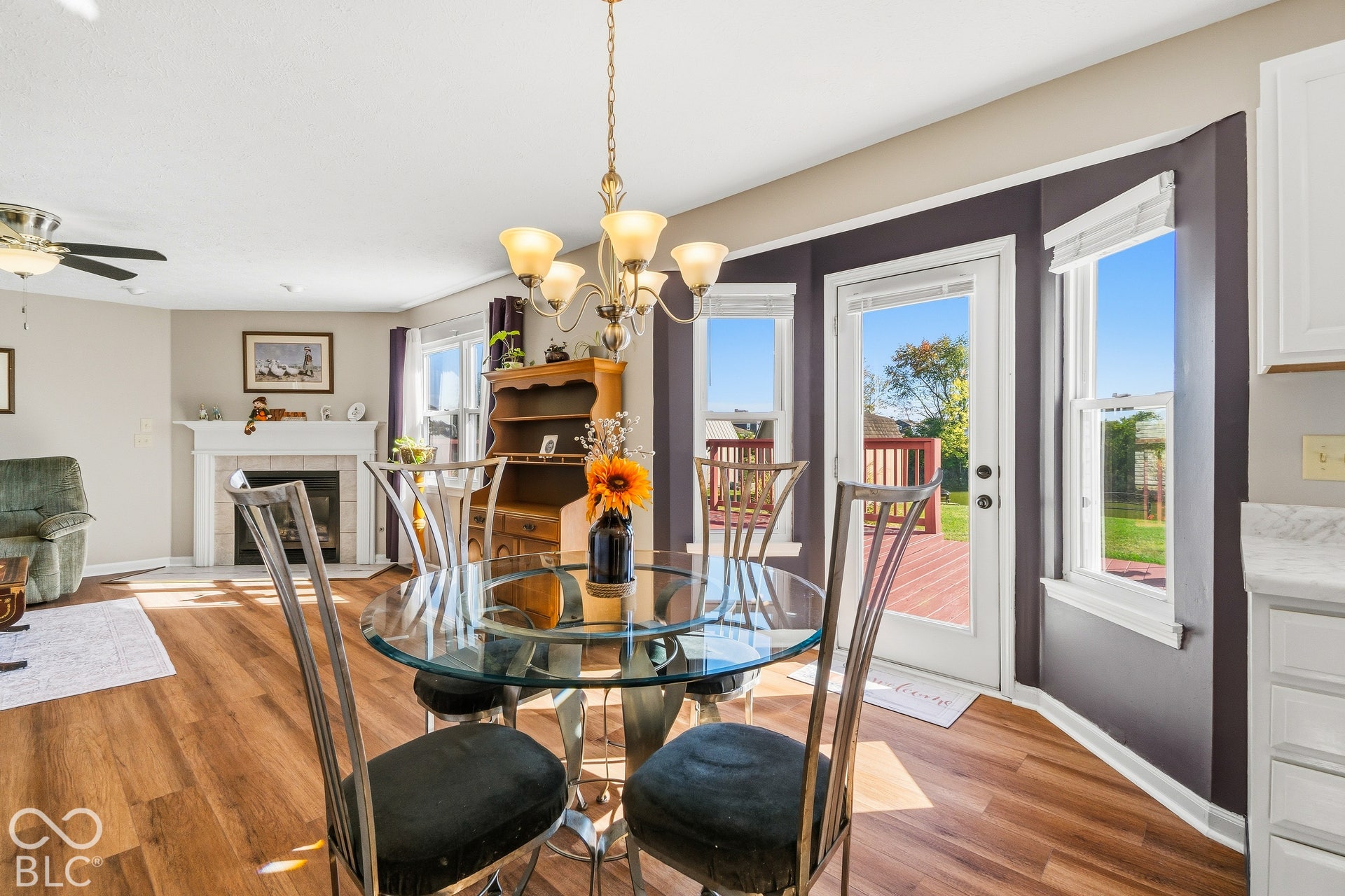 Wood-finish flooring throughout, chandelier lighting, fireplace with mantel and tile surround, white door leading to an outdoor deck, and bay windows