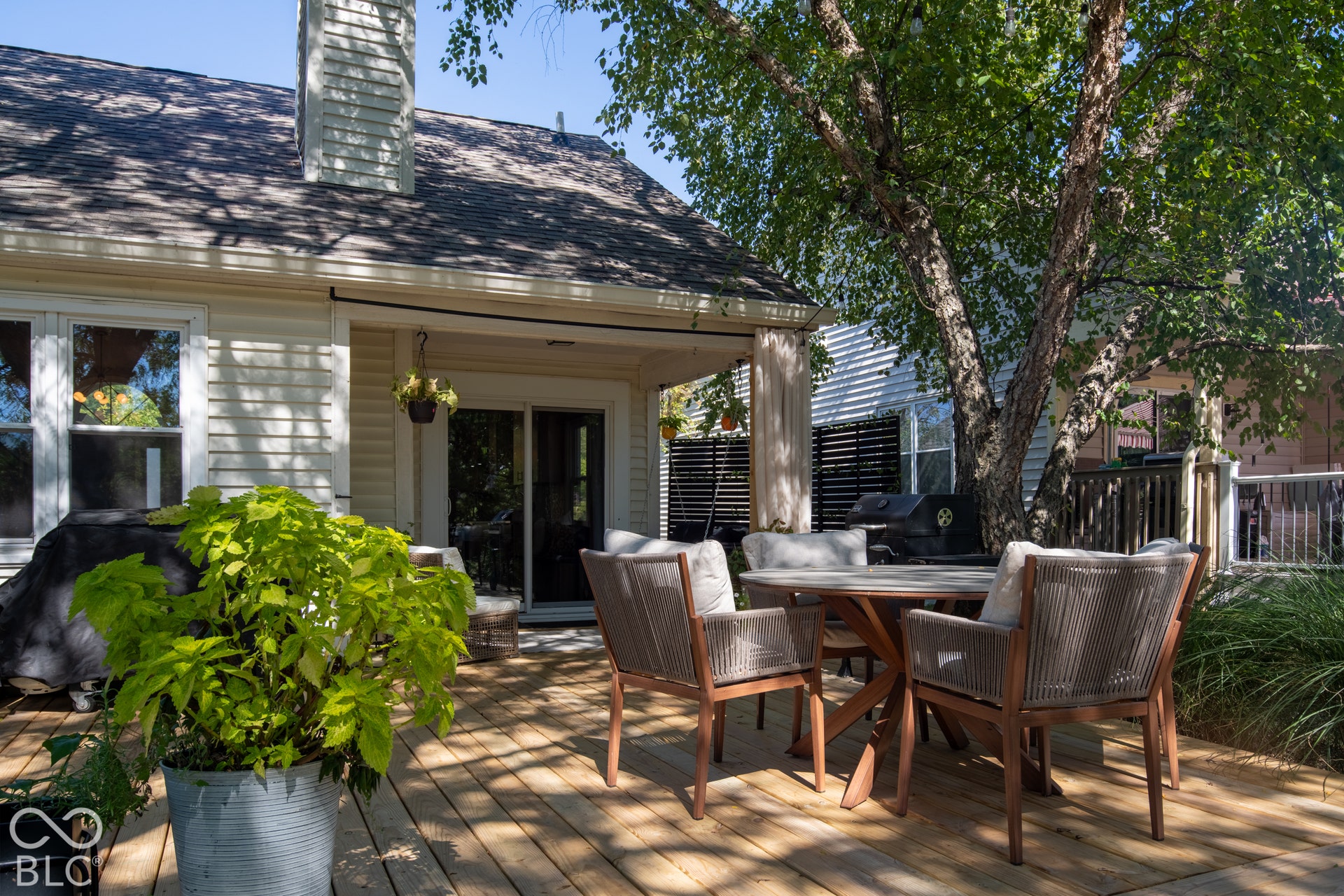 Expansive wood-finish deck with a mature shade tree, featuring a sliding glass door and a covered porch with a privacy screen