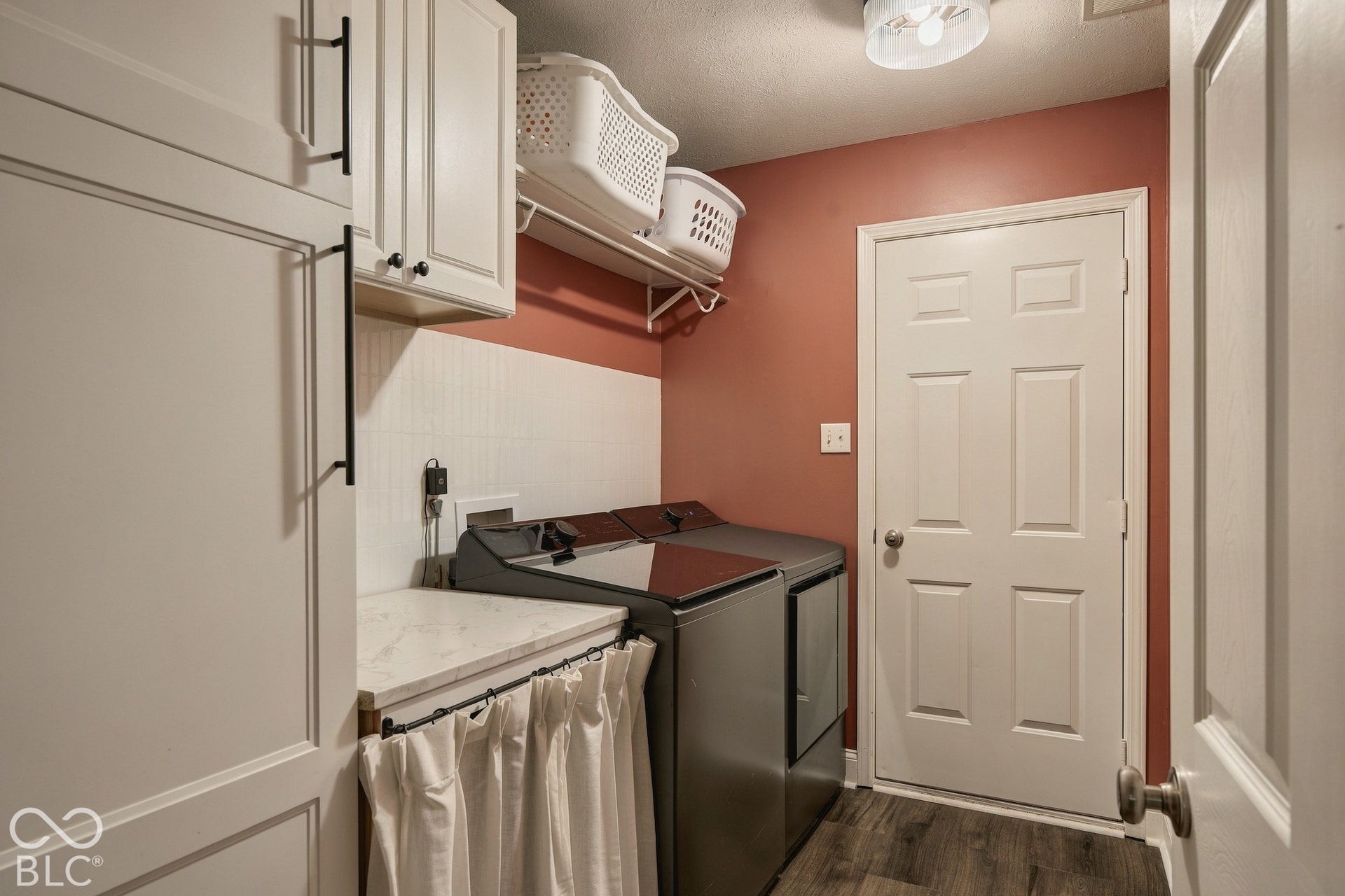 Laundry area featuring wood-finish flooring, built-in white cabinetry with matte black hardware, a light-toned countertop, and a white tile backsplash