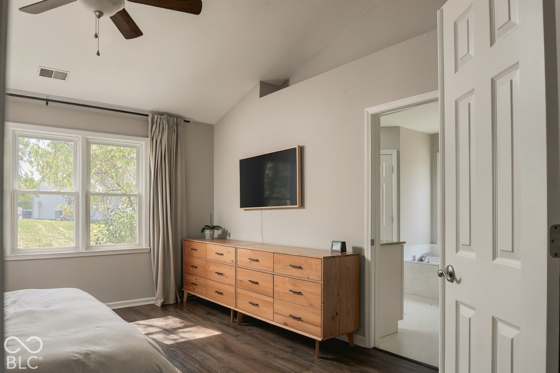 Cathedral ceiling with a ceiling fan, dual windows with exterior views, dark wood-finish flooring, and an adjoining bathroom with a built-in tub