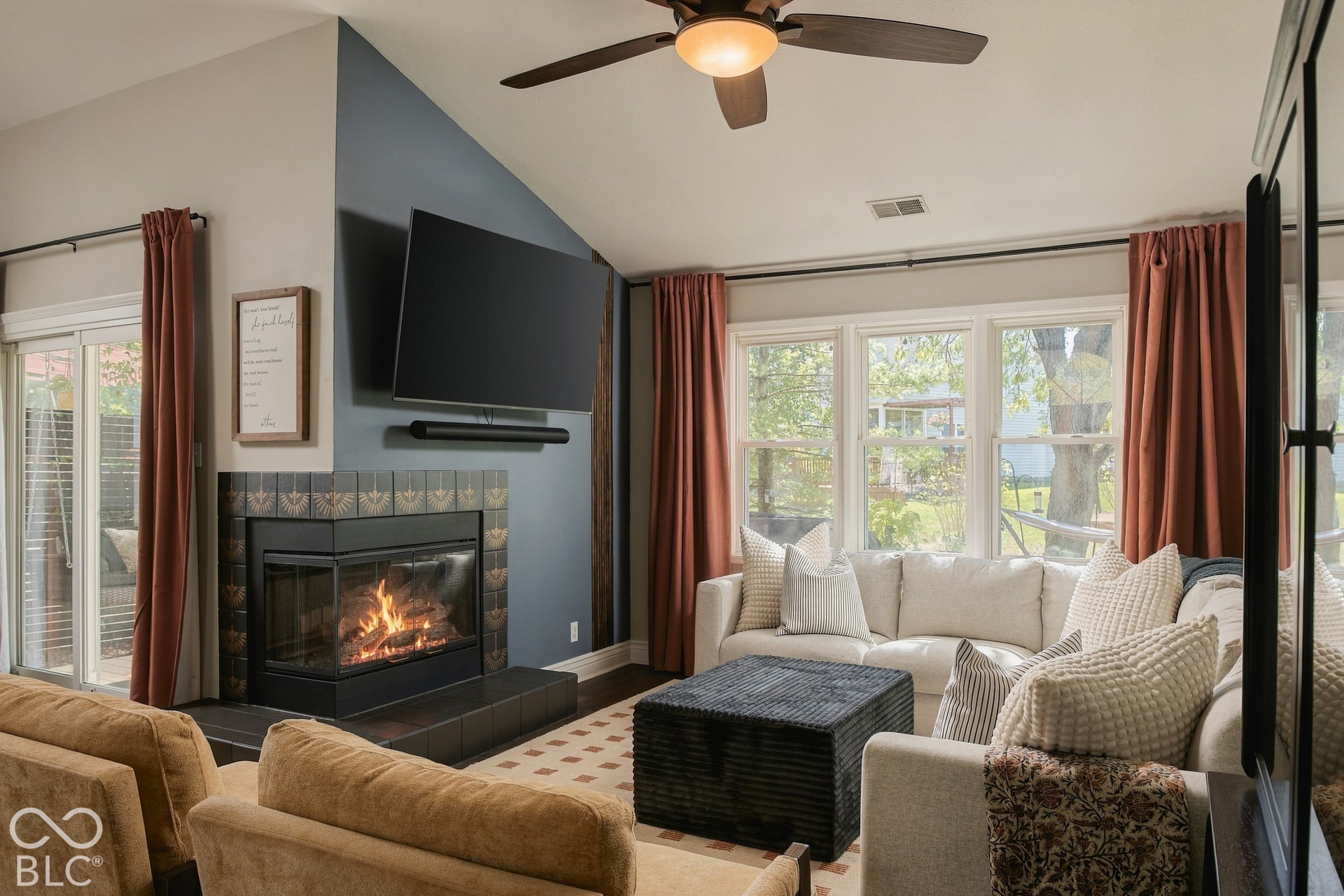 Vaulted ceiling with a ceiling fan, corner fireplace with a tiled surround, large window bank, and wood-finish flooring