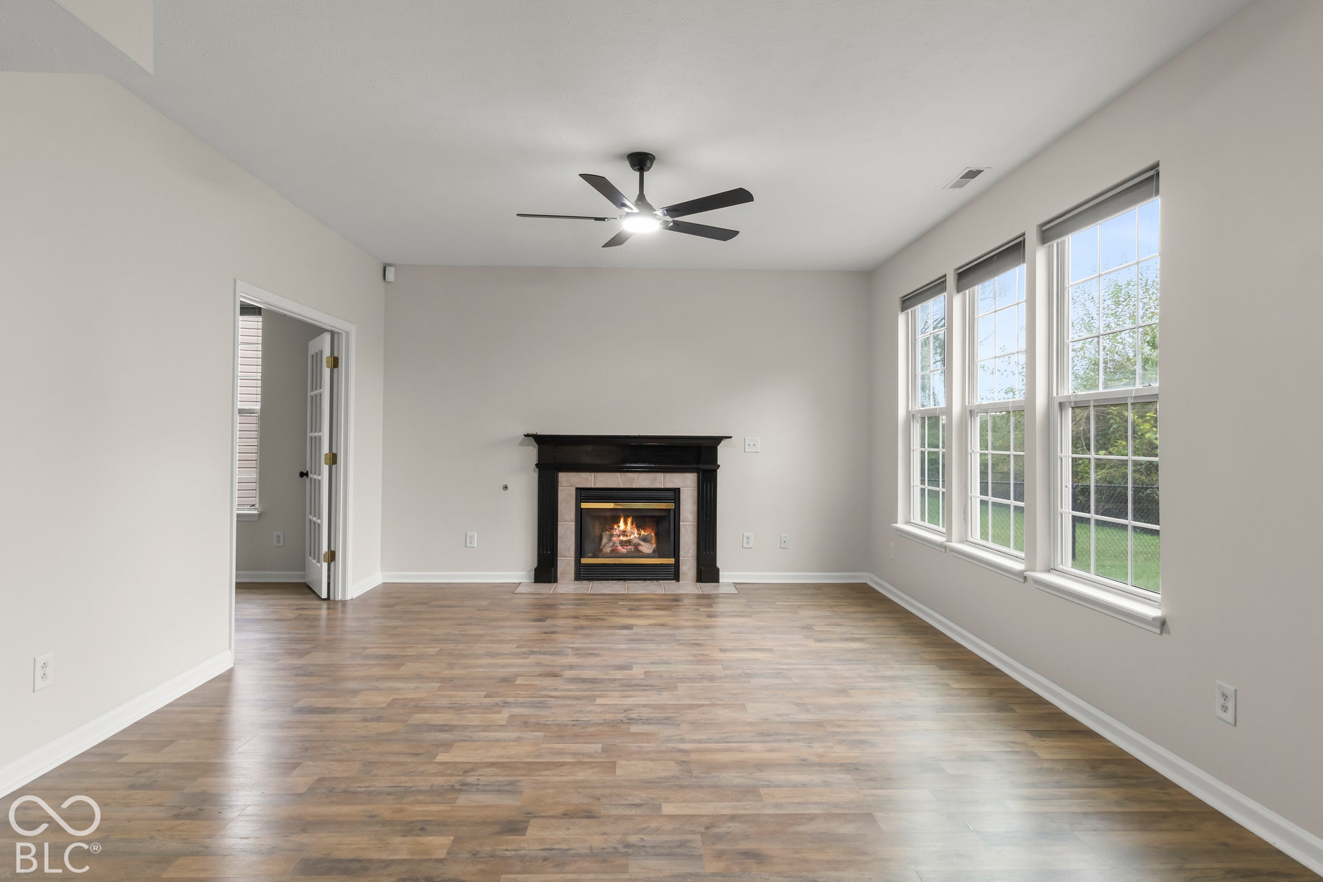 Wood-finish flooring, a ceiling fan, a fireplace with a dark mantel, and multiple windows with blinds