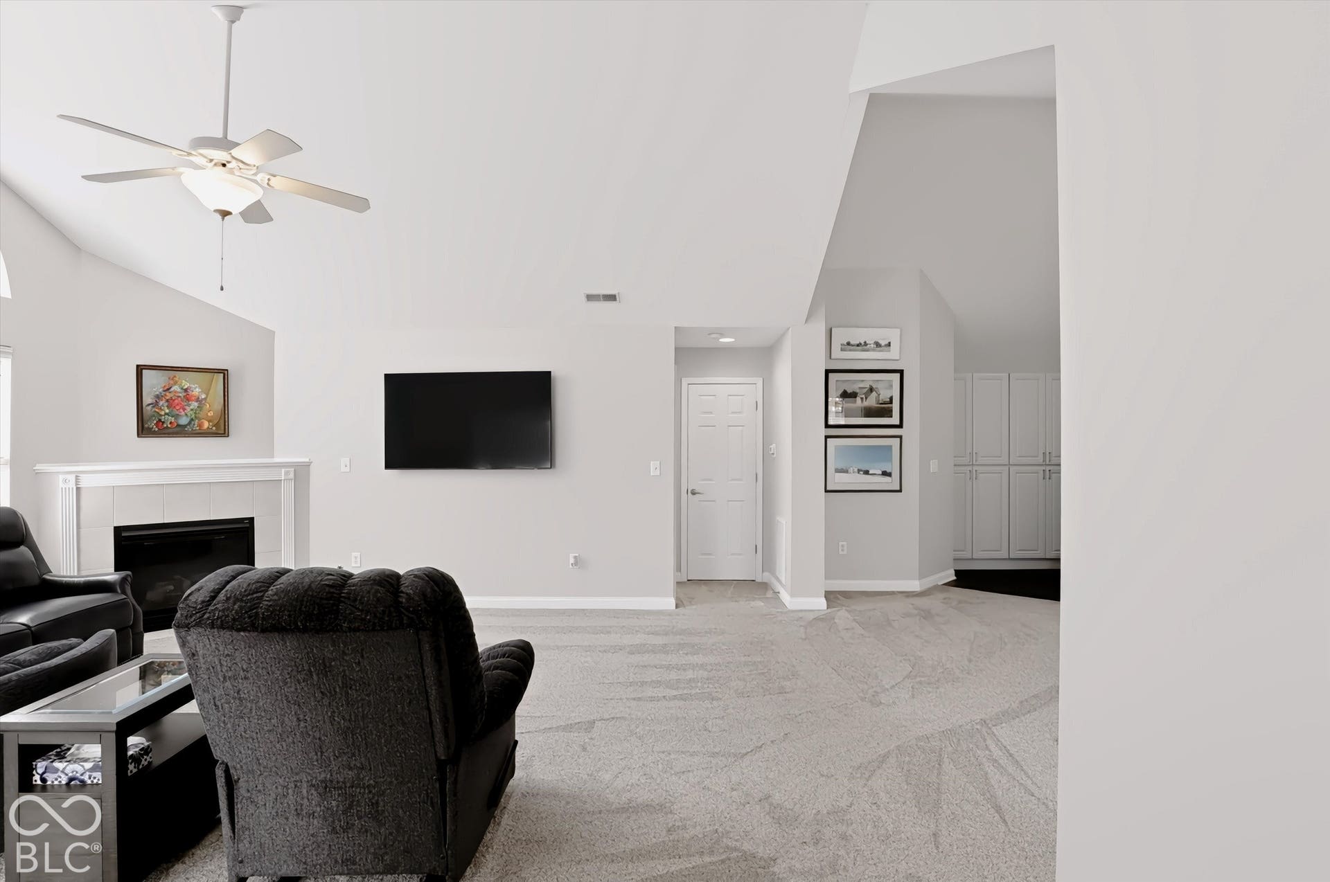 Vaulted ceiling with ceiling fan, white mantel over a dark fireplace, light gray wall paint, light-toned carpet flooring, and white built-in cabinetry