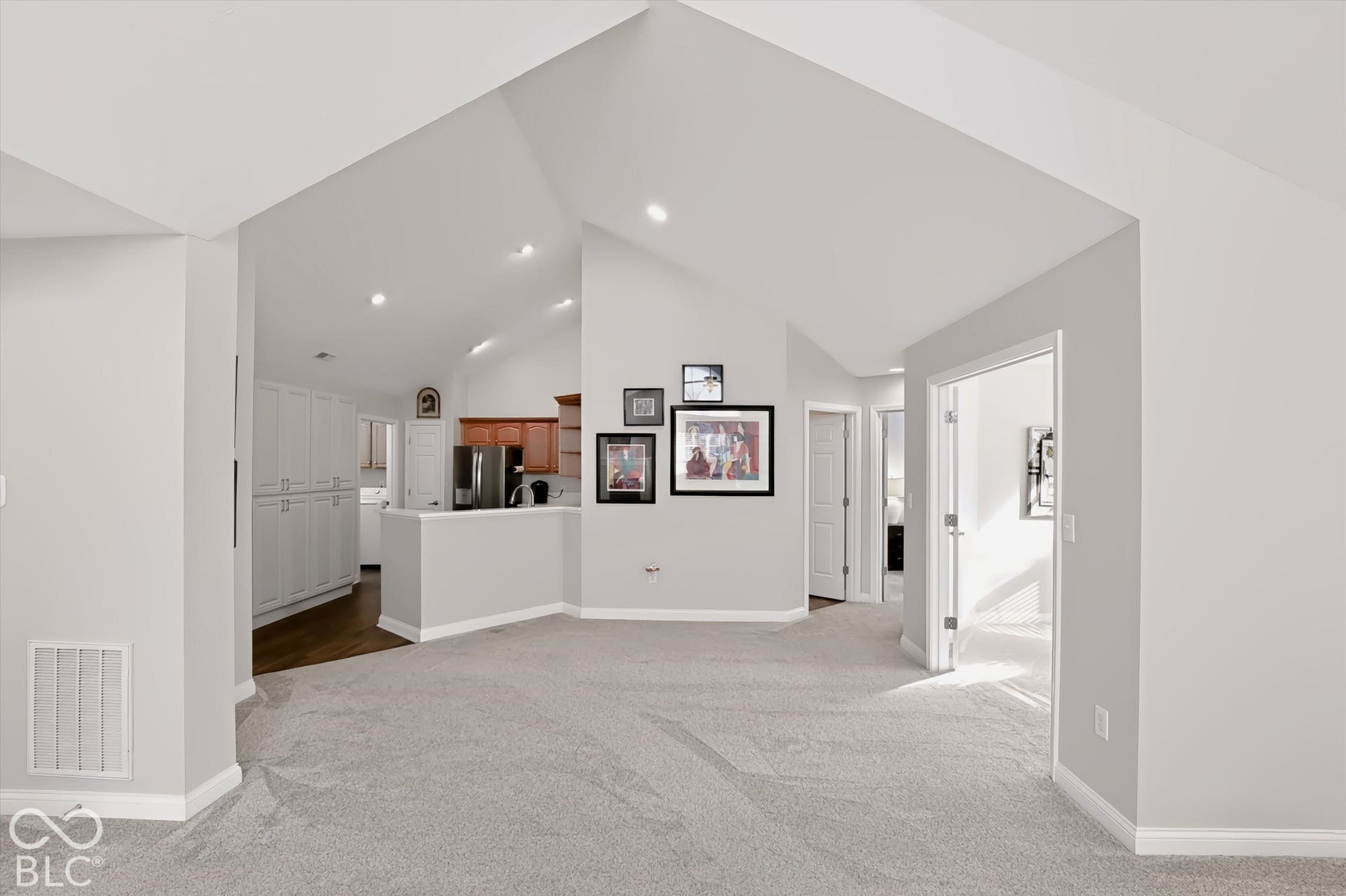 Vaulted ceilings with recessed lighting, light-toned carpeting, white trim, and a partial view of the kitchen with wood-finish cabinetry