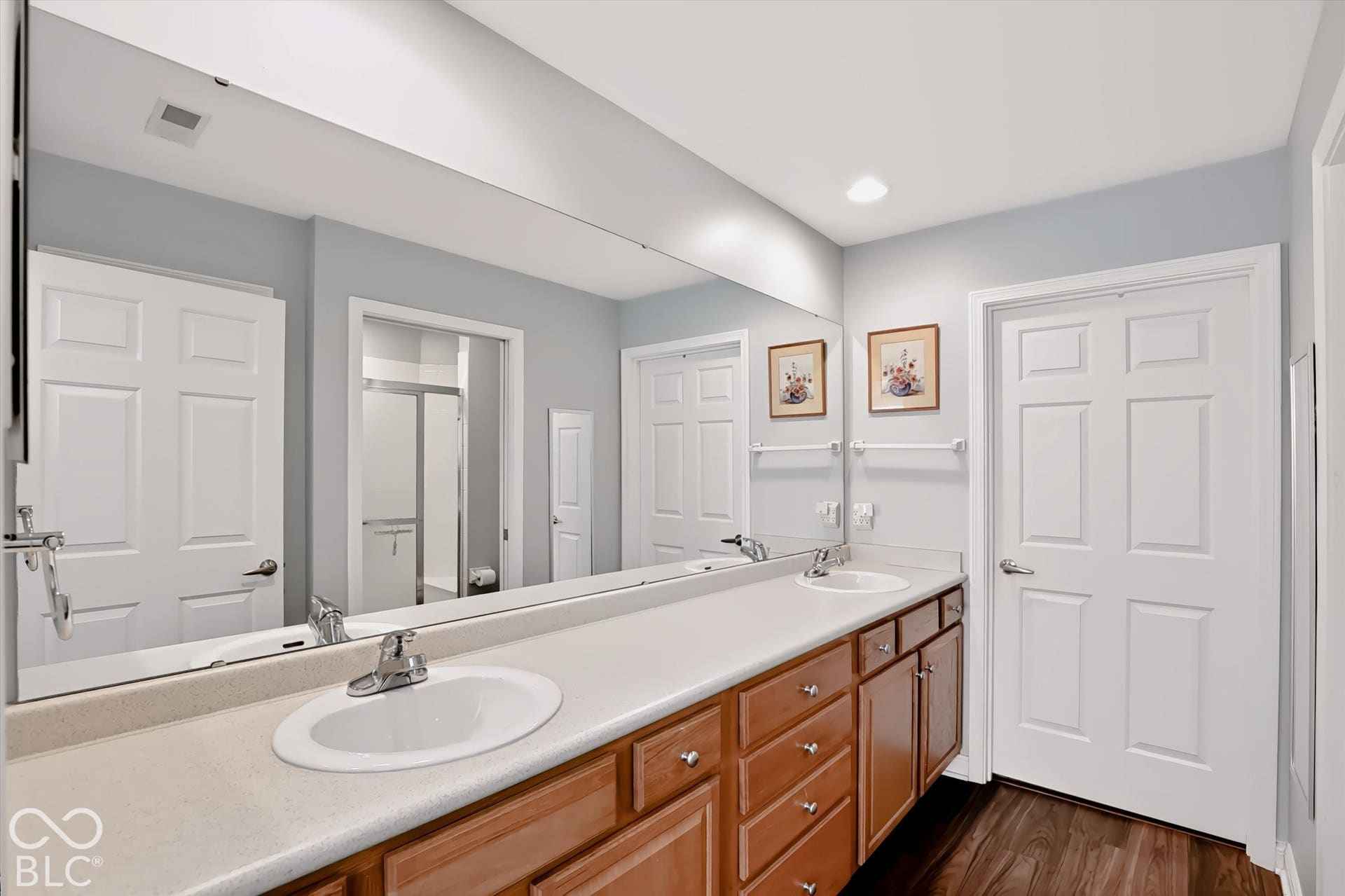 Dual vanity with integrated sinks, expansive wall-mounted mirror, wood-finish flooring, and recessed lighting