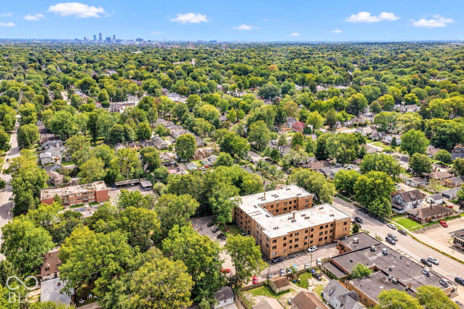 Urban landscape featuring a large multi-story building, surrounded by abundant mature trees and residential structures