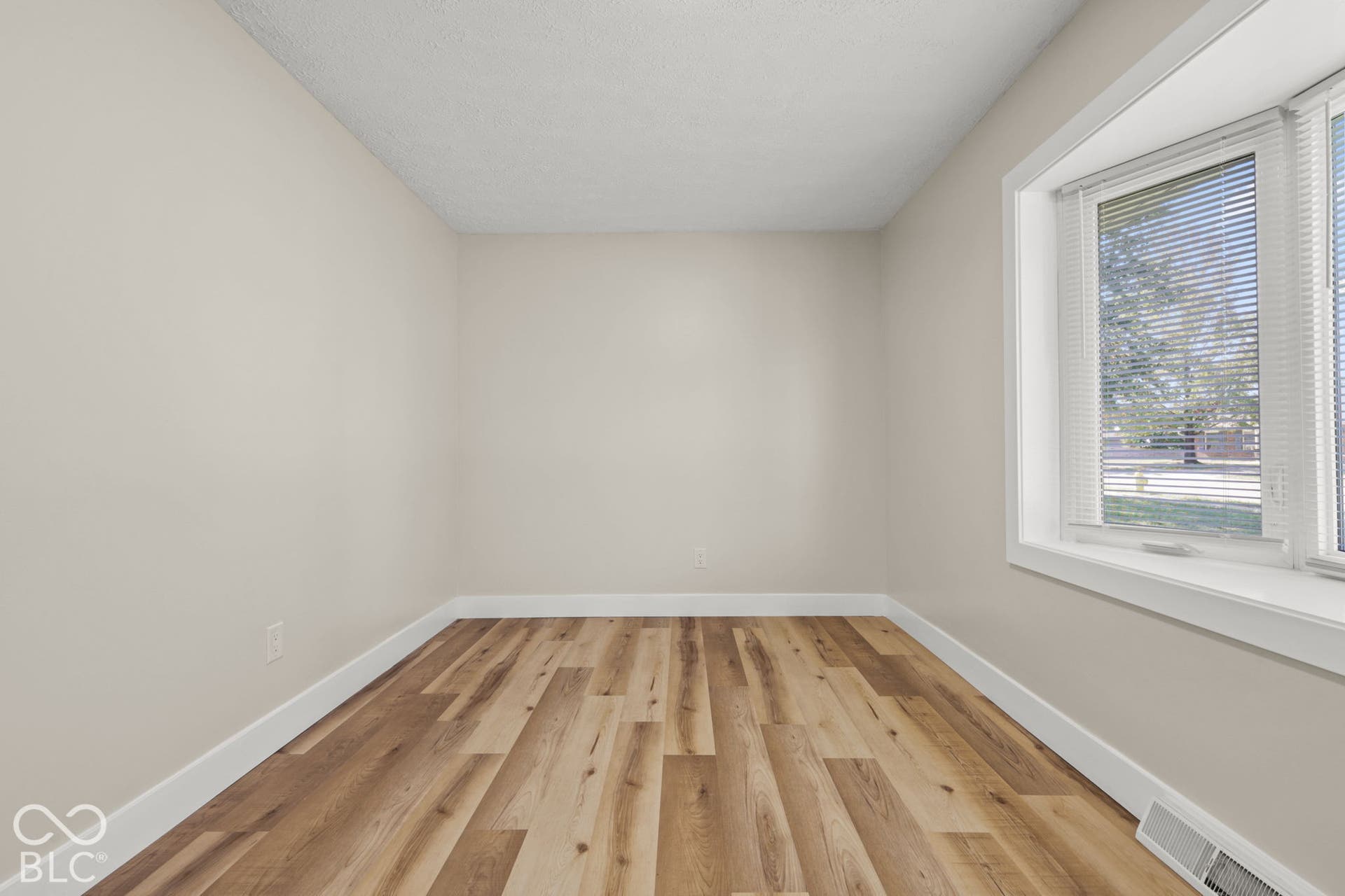 Light-toned walls with wood-finish flooring, featuring a window with blinds and white trim