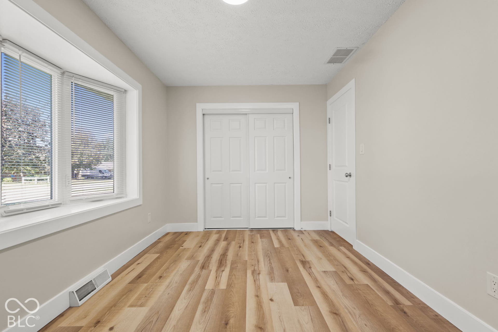 Bay window with white blinds, wood-finish flooring, white bi-fold closet doors, white wall paint, and overhead lighting