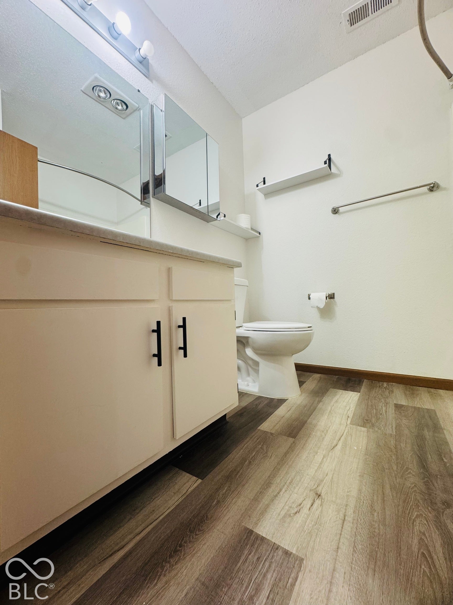 Light wood-finish flooring, white vanity with black hardware, white countertop, and wall-mounted mirror with overhead lighting