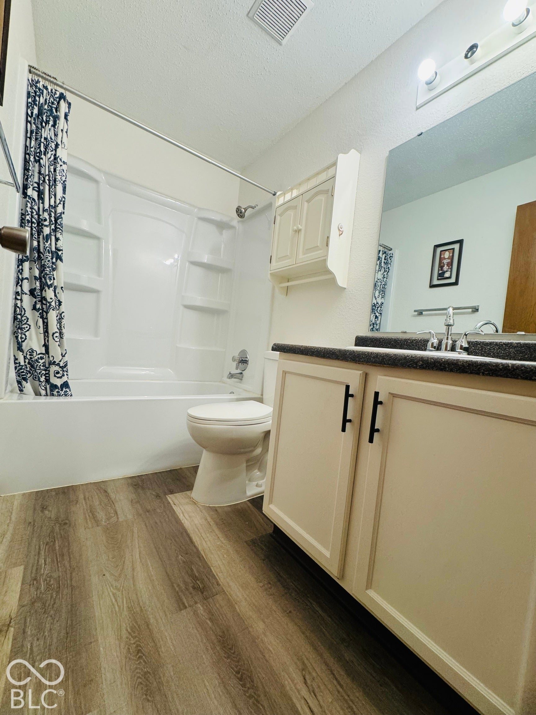 Wood-finish flooring, white vanity with black hardware, dark countertop, recessed wall cabinet, and a white tub and shower combination