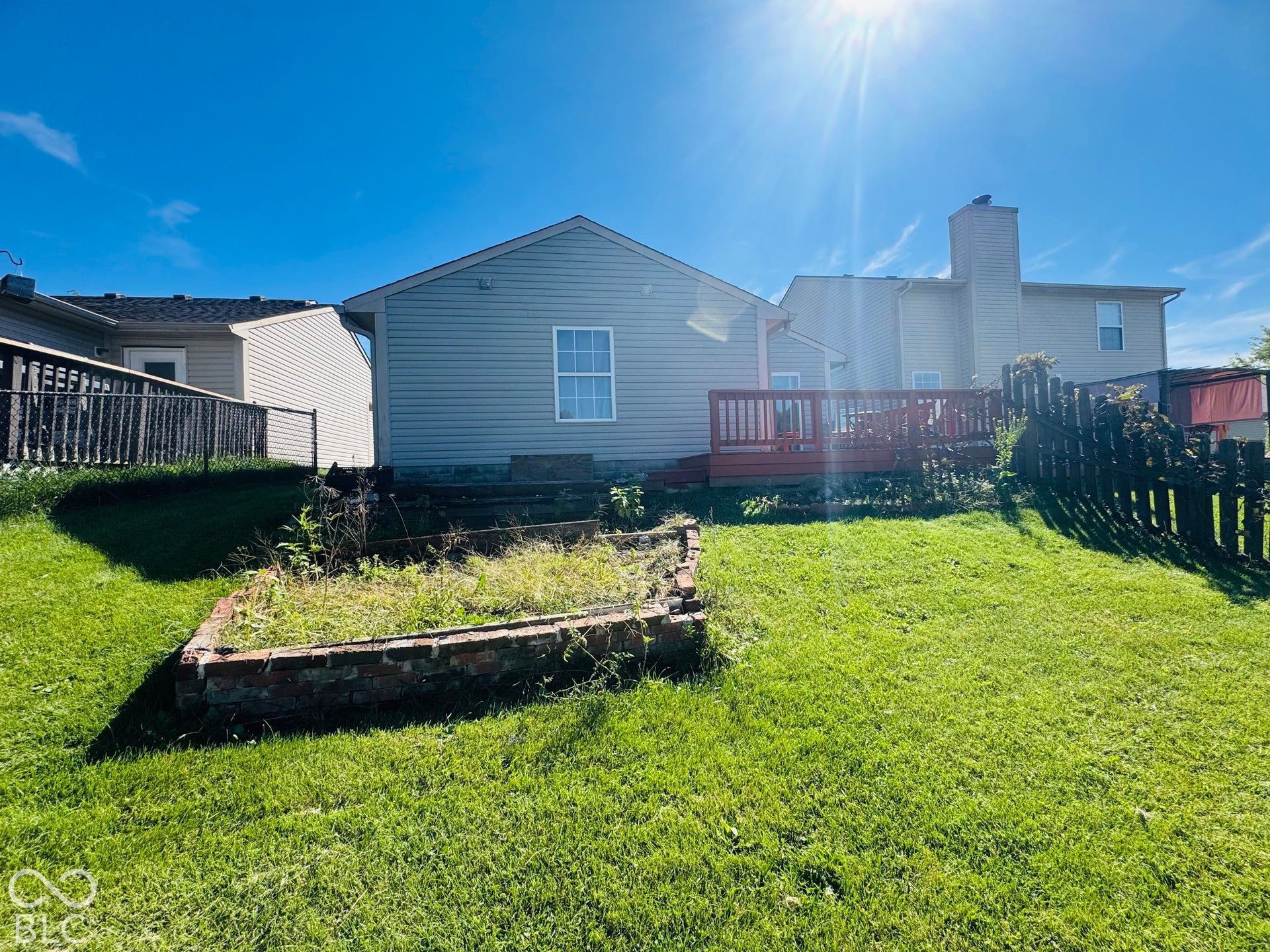 Expansive green lawn featuring a red-stained wood deck, brick-lined garden bed, and a partial wood fence