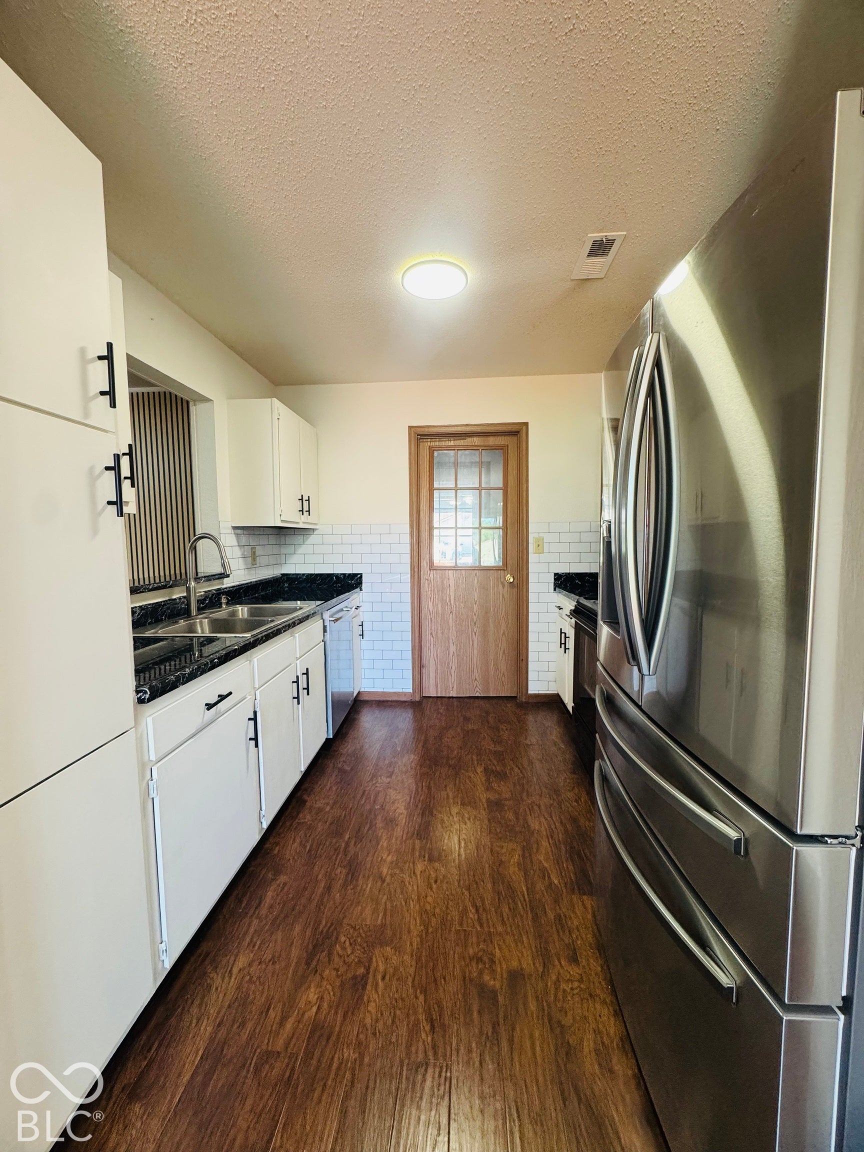 Galley kitchen featuring wood-finish flooring, white cabinetry with black hardware, dark countertops, white subway tile backsplash, and a stainless steel refrigerator