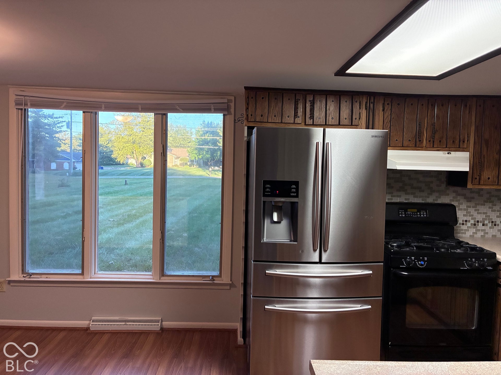 Stainless steel refrigerator, black range, wood-finish flooring, mosaic tile backsplash, and a large window