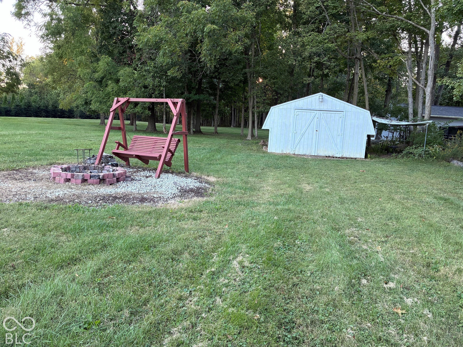 Expansive grassy outdoor space featuring a fire pit with brick surround, a wooden swing, and a white storage shed