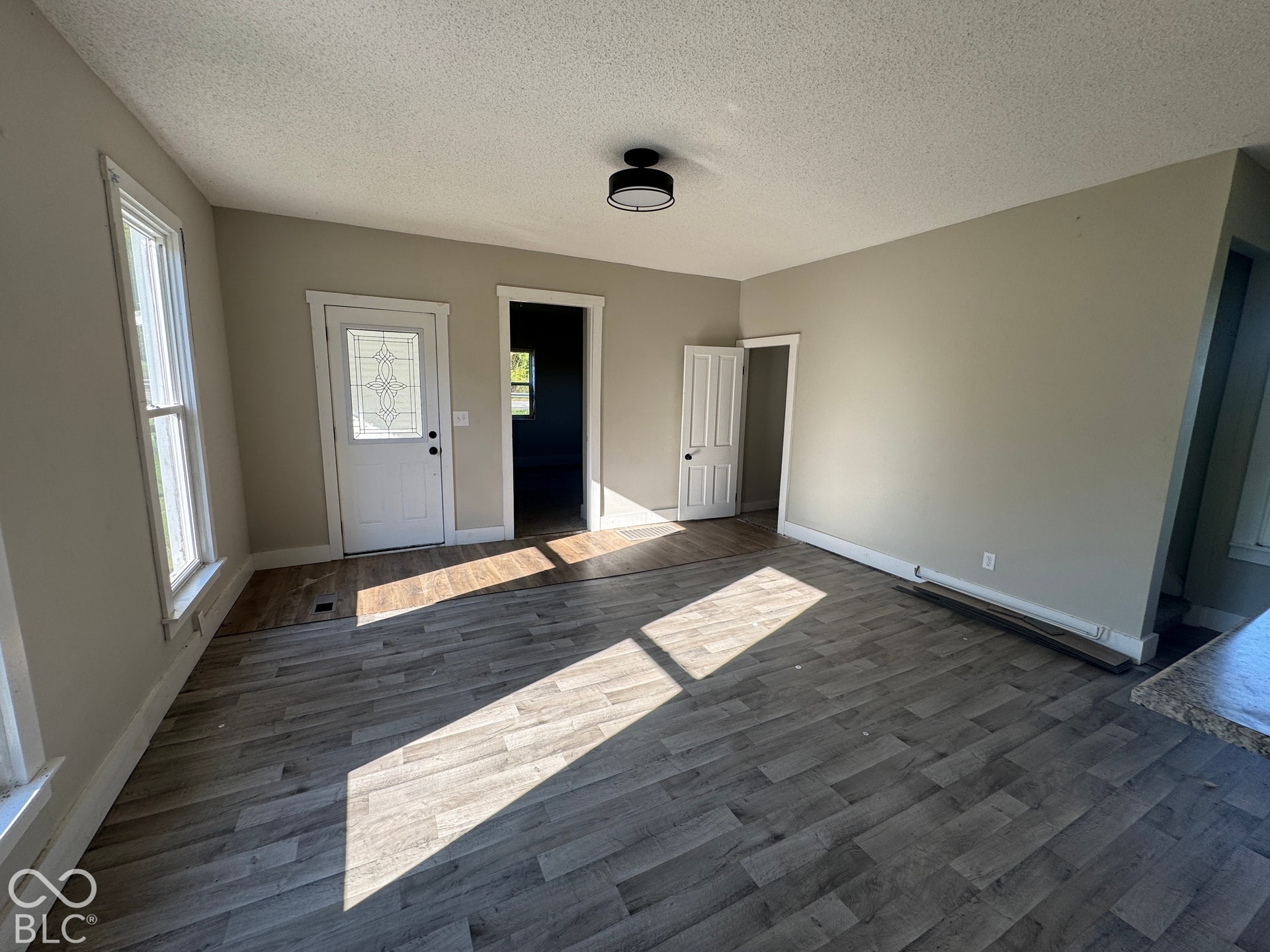 Spacious room featuring wood-finish flooring, neutral-toned walls, a decorative glass panel door, multiple windows, and a flush mount light fixture