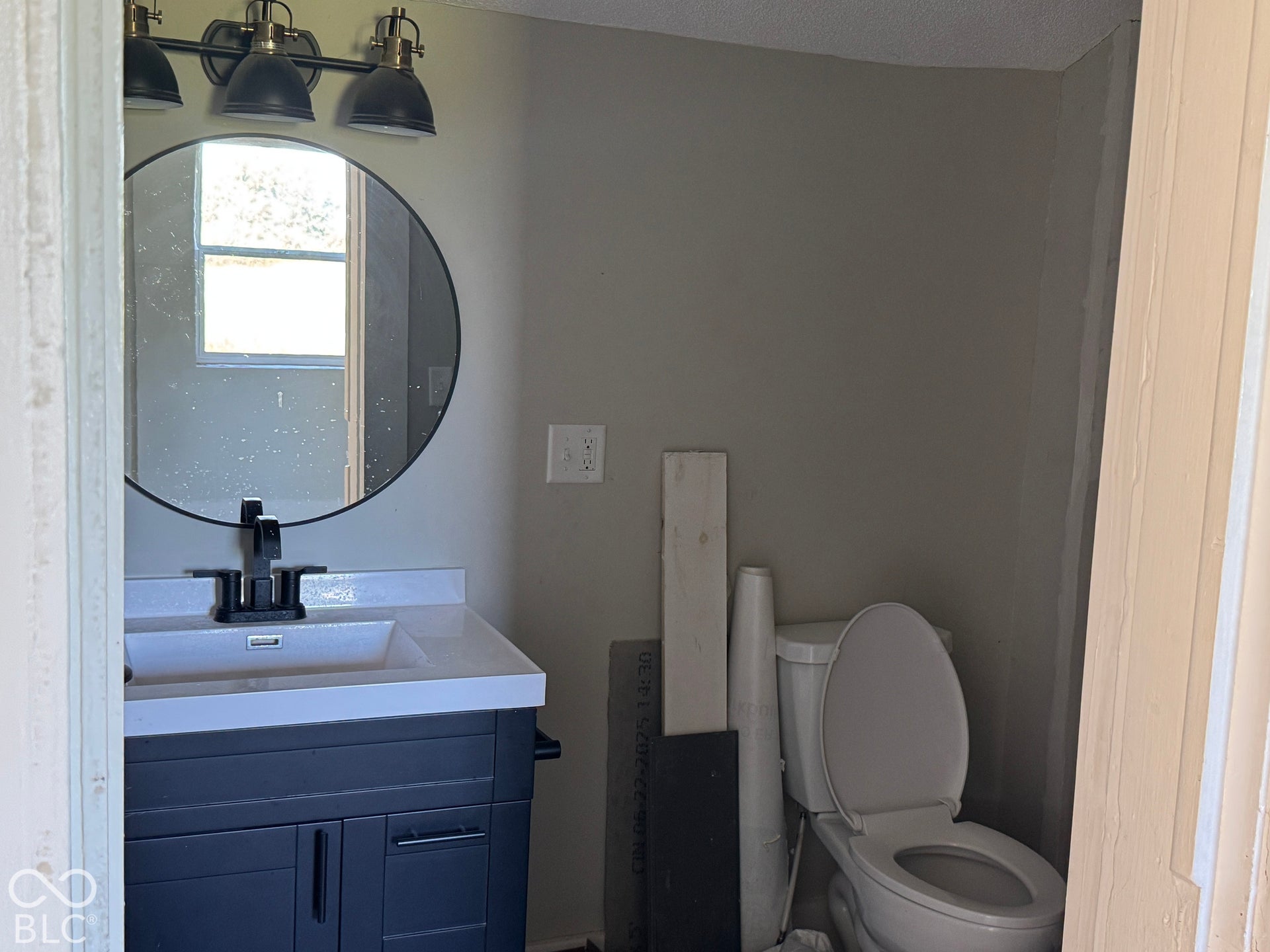 Vanity with a white countertop and integrated sink, black wall-mounted faucet, round mirror, and three-light fixture