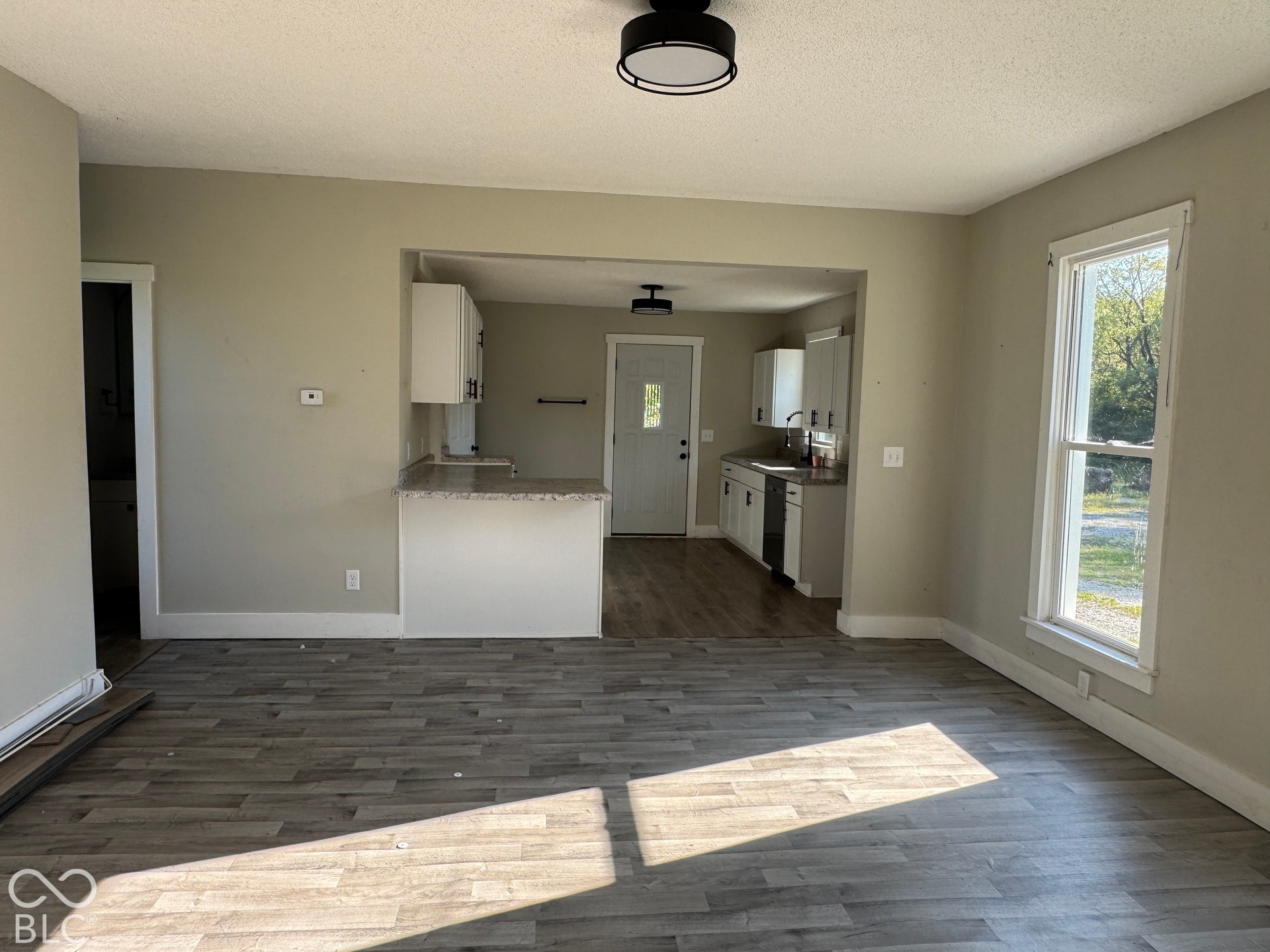 Wood-finish flooring throughout, light-colored walls, recessed kitchen area, white cabinetry, and a double-hung window