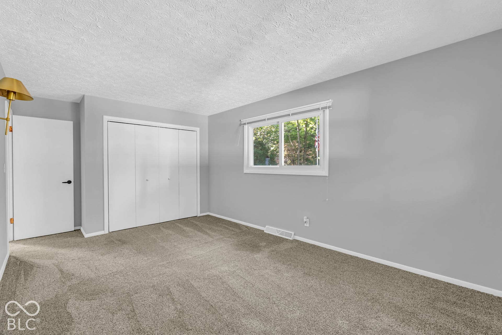 Textured ceiling, light gray walls, window with white trim, gray carpet flooring, and white bi-fold closet doors