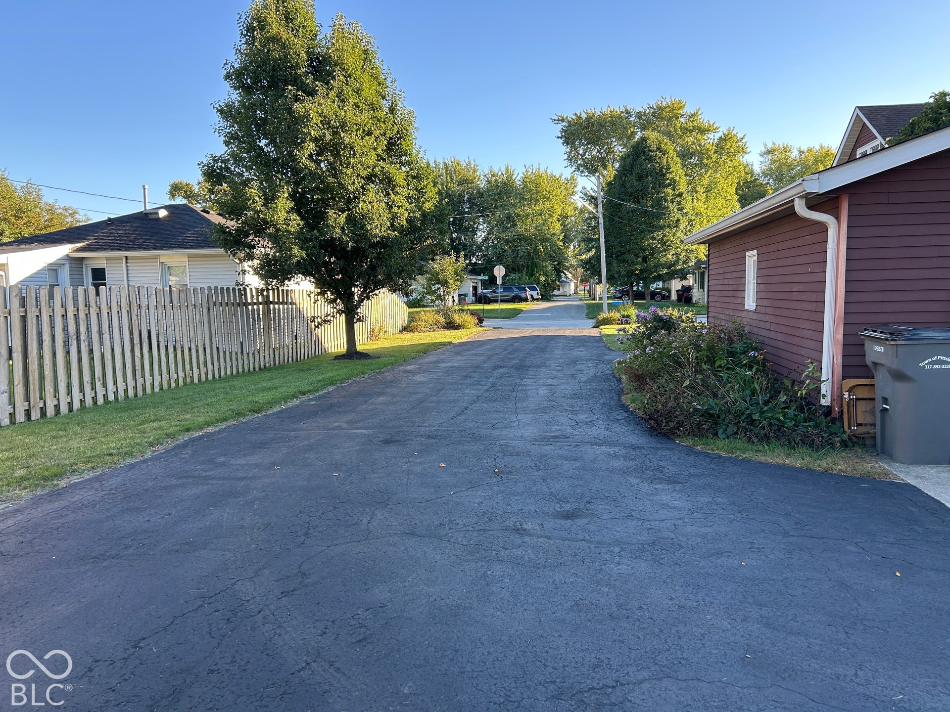 Paved driveway leading to a detached structure with siding, surrounded by mature trees and a wooden fence