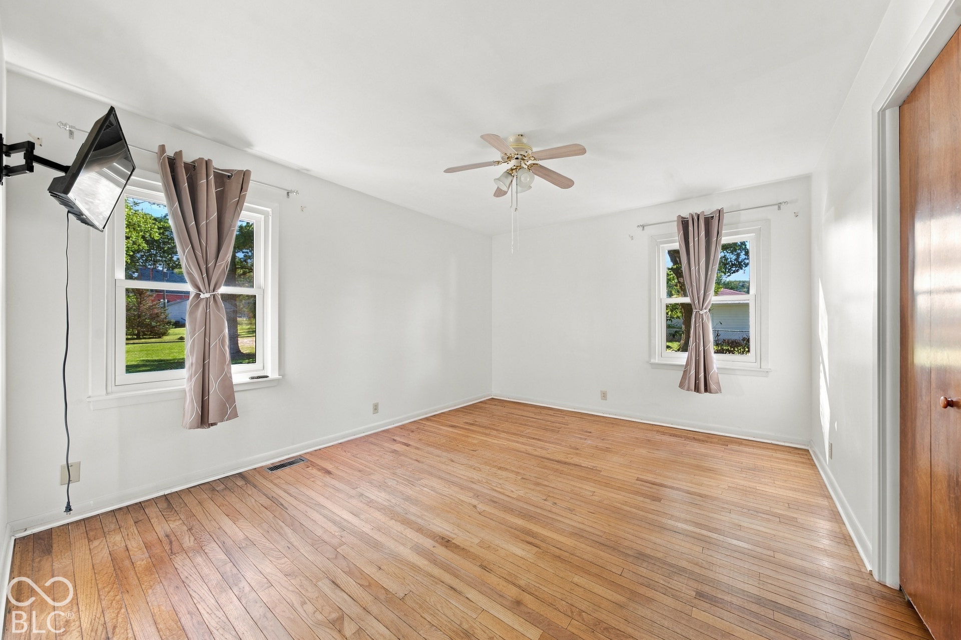 Hardwood flooring throughout, two windows, ceiling fan with integrated lighting, and a wood-paneled door