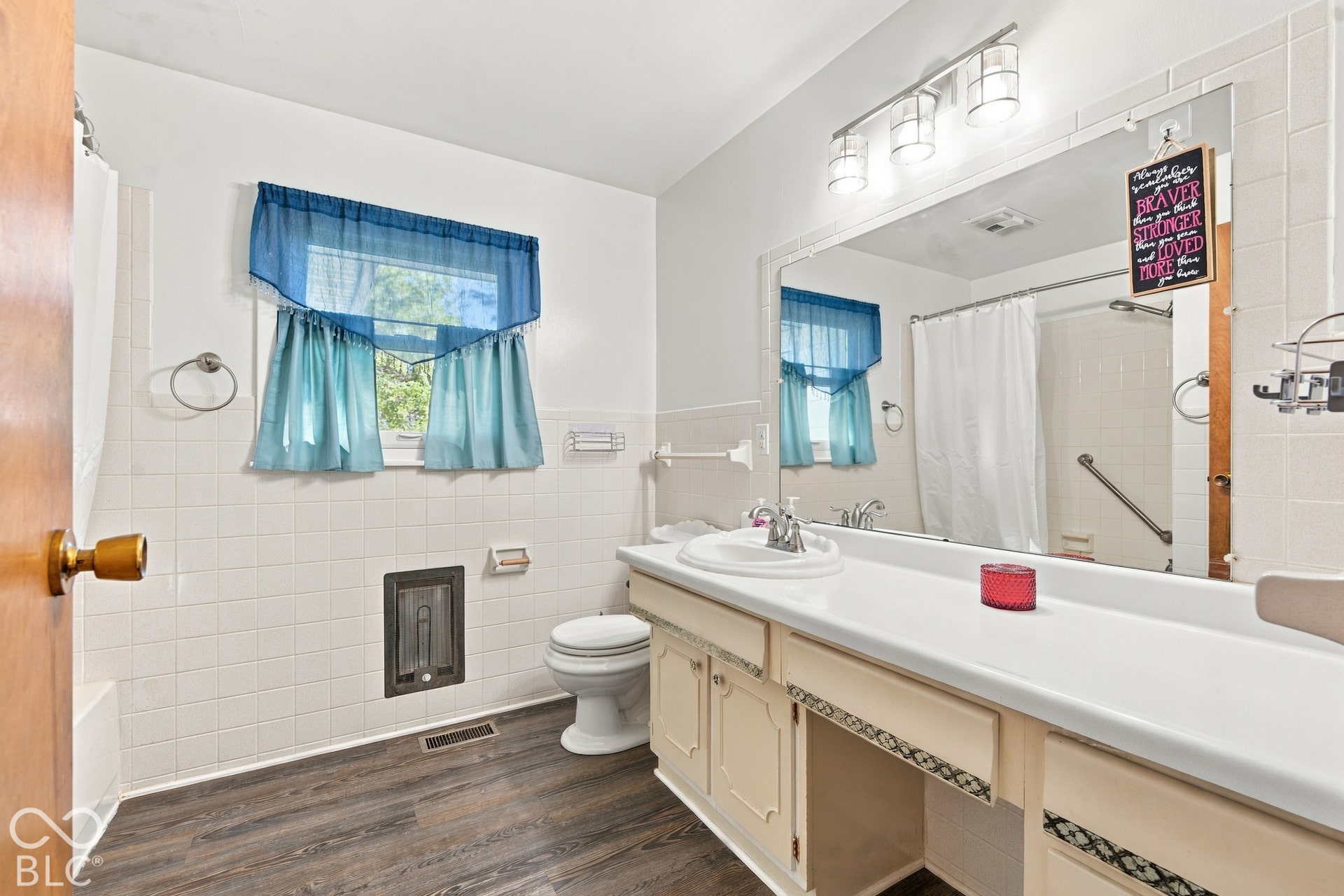 Bathroom featuring a single vanity with an integrated sink, broad wall mirror, wood-finish flooring, white tile walls, and a window