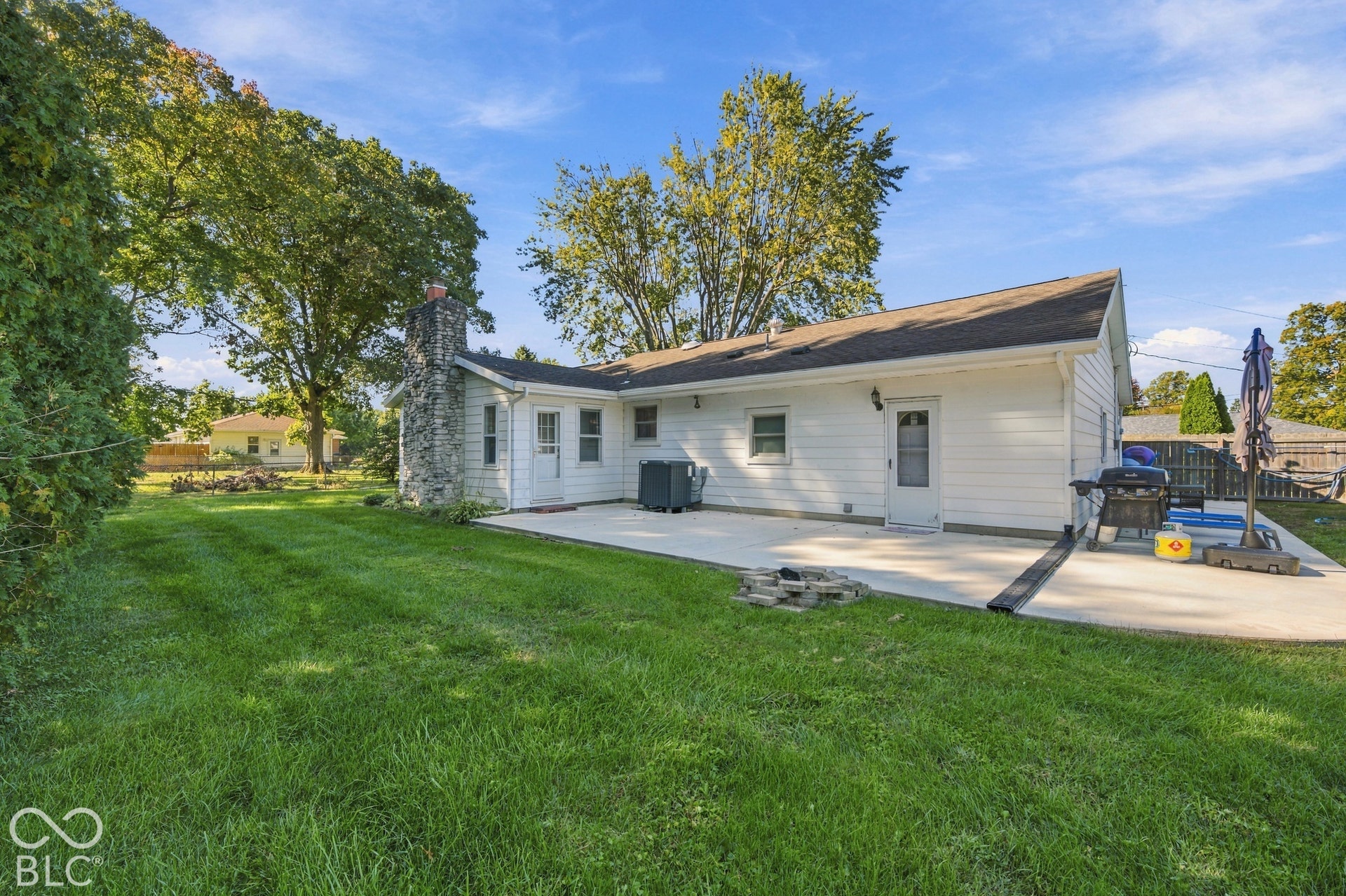Expansive grass lawn, concrete patio, white siding, stone chimney, and mature trees