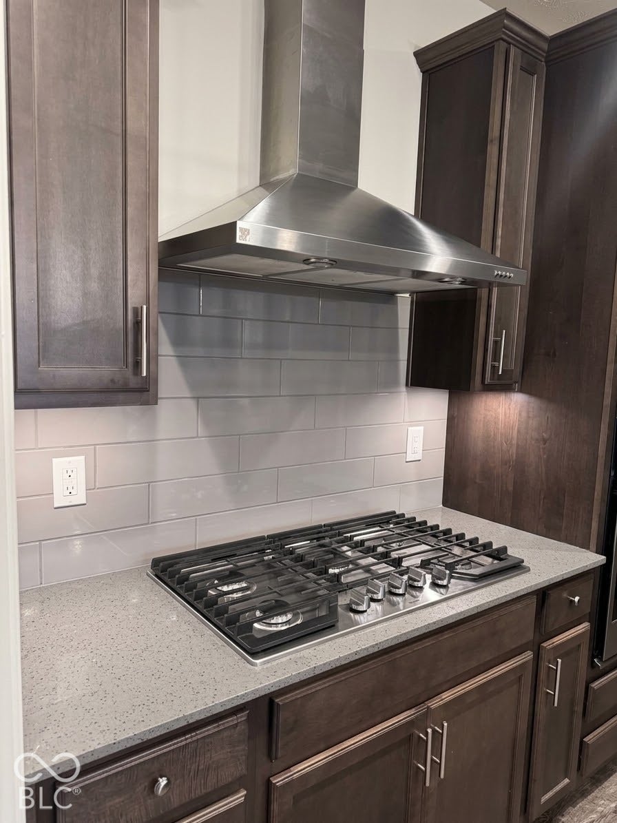 Stainless steel range hood with a gas cooktop, light grey subway tile backsplash, and dark wood-finish cabinetry