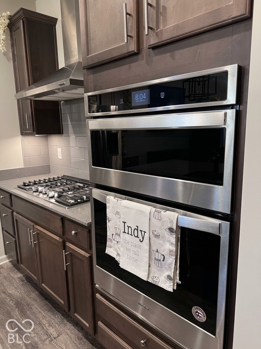 Built-in stainless steel double oven, gas cooktop with stainless steel range hood, dark wood-finish cabinetry, light gray countertop, and subway tile backsplash