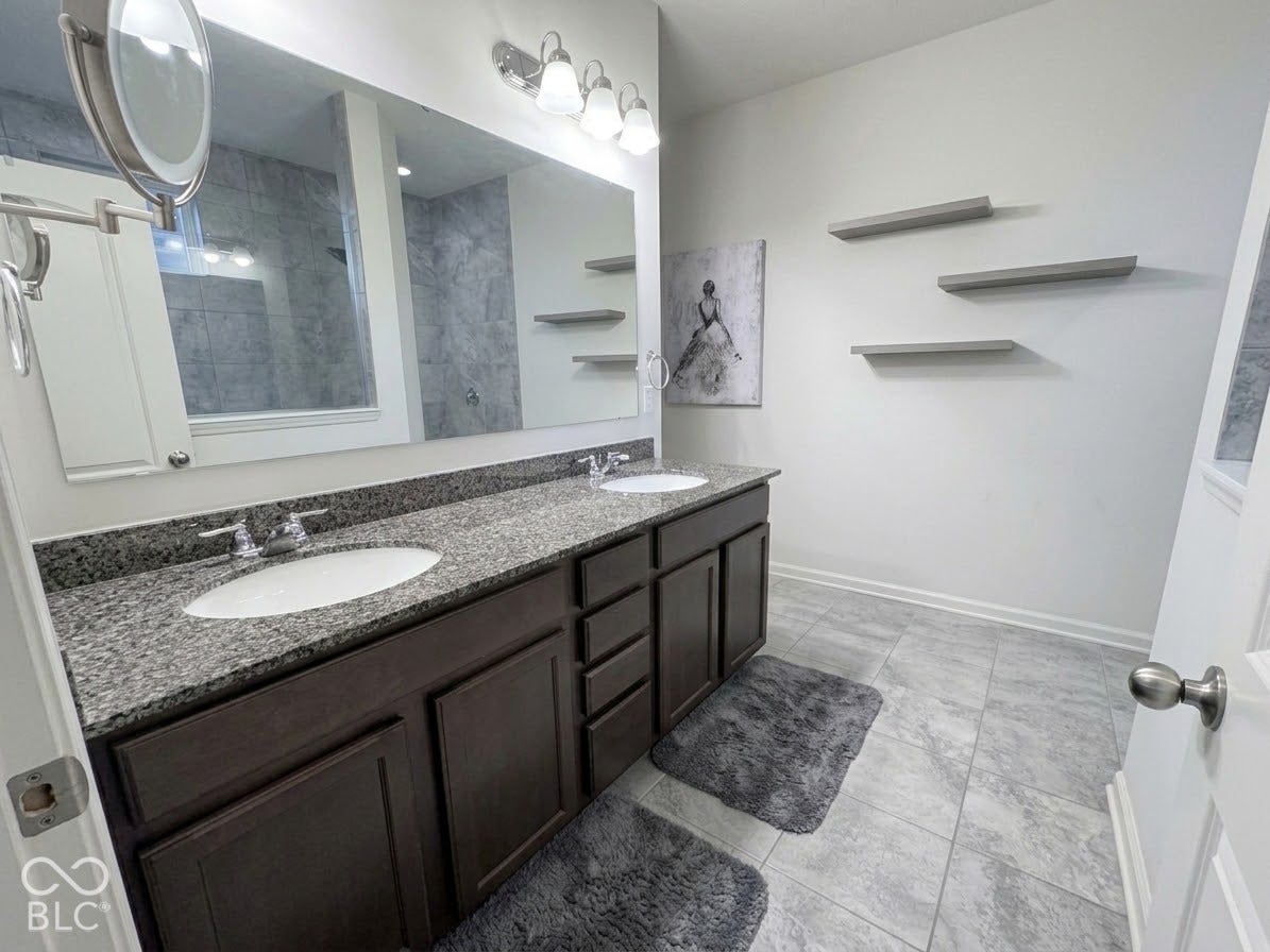 Dual vanity with granite countertop, integrated sinks, dark wood-finish cabinetry, ceramic tile flooring, and a large wall-mounted mirror