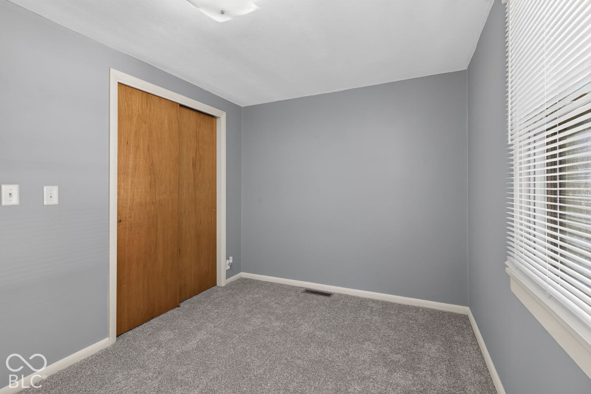 Gray wall finish, light gray carpet flooring, wood-finish sliding closet doors, white window trim, and a flush-mount ceiling light fixture