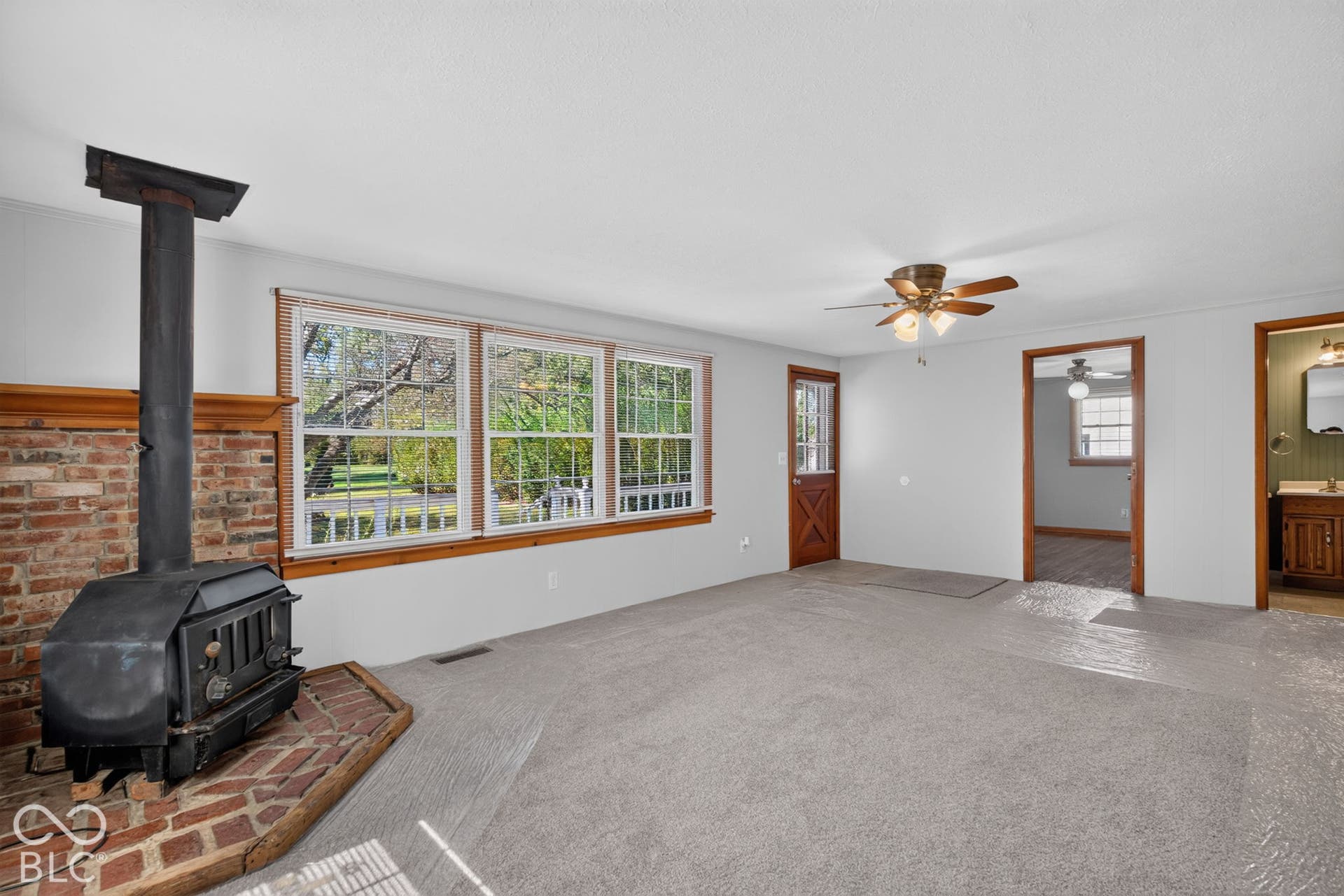 Brick accent wall with wood stove, three large windows, light gray carpeting, wood-finish ceiling fan, and wood-paneled door