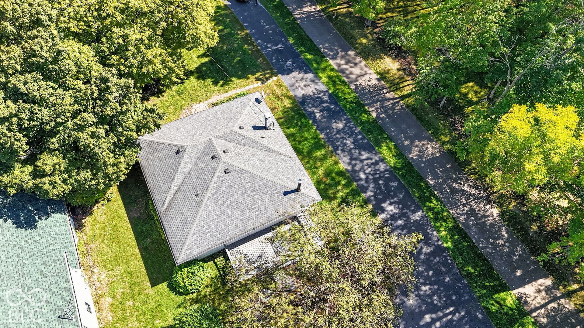Gable roofline with asphalt shingles, mature surrounding trees, and a paved walkway
