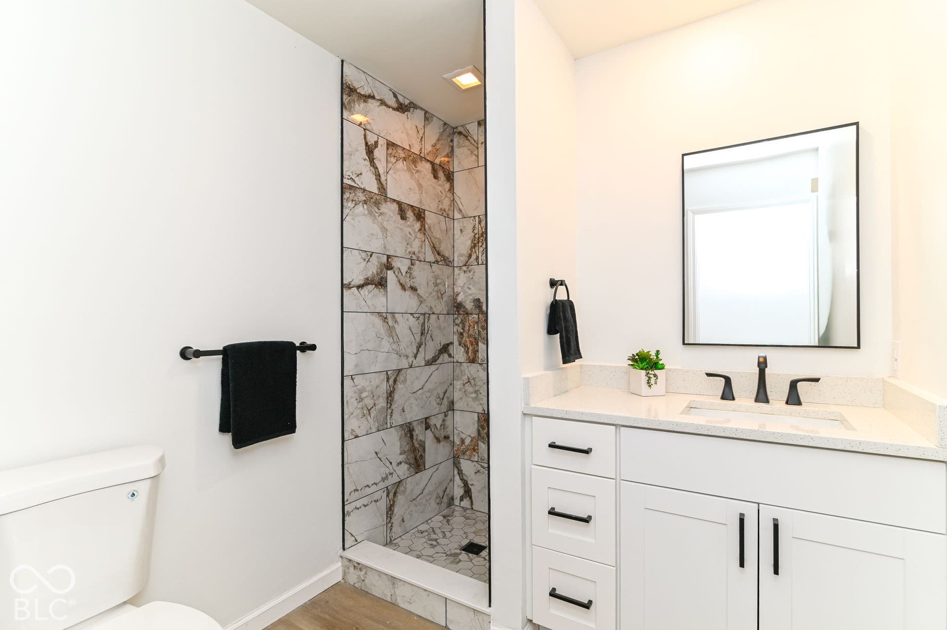 Walk-in shower with marble-look tile surround, white vanity with integrated sink, quartz countertop, black hardware, and rectangular framed mirror
