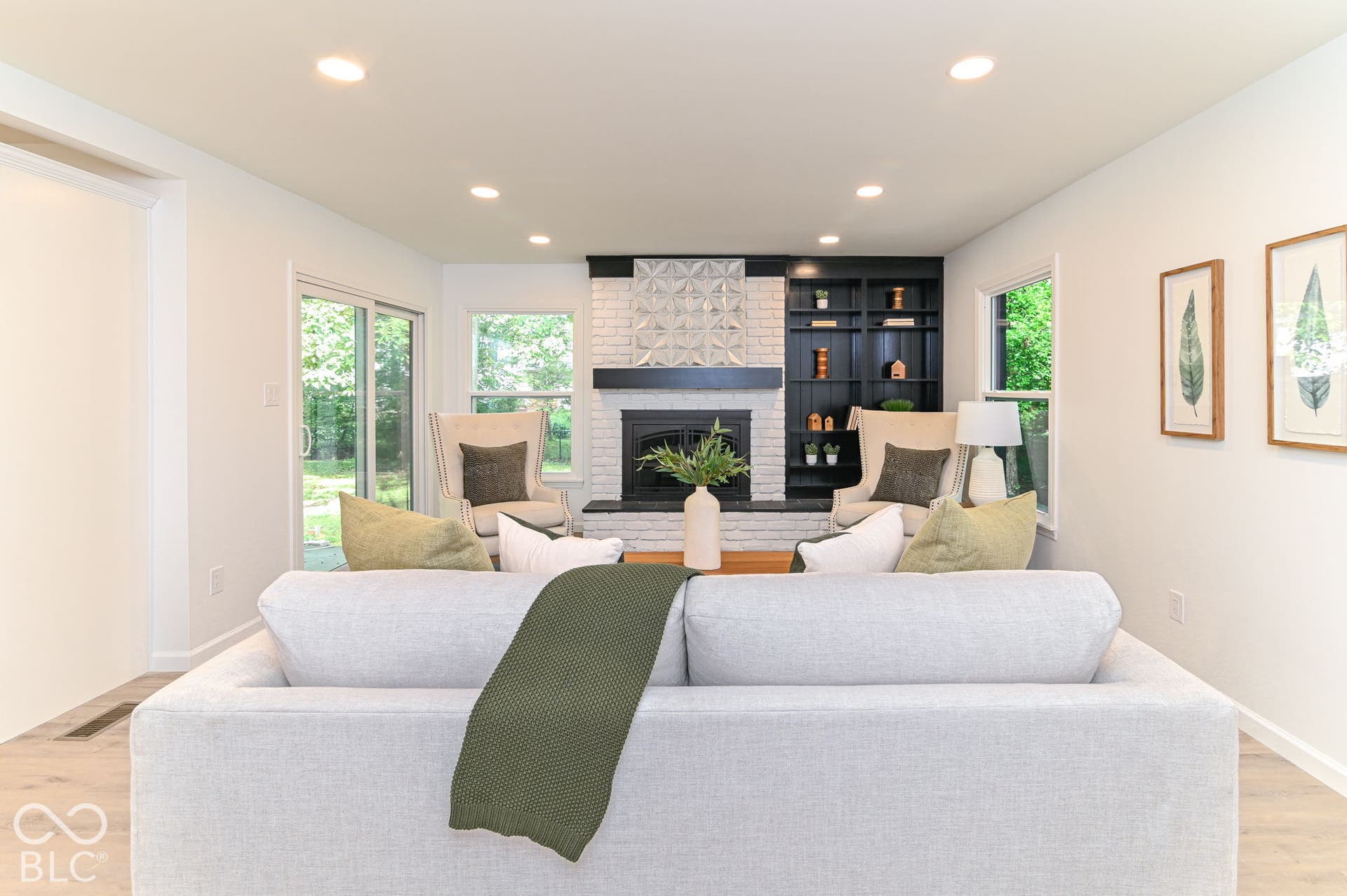 Recessed lighting illuminating the bright interior, featuring a white brick fireplace with a dark wood mantel, built-in shelving, and wood-finish flooring