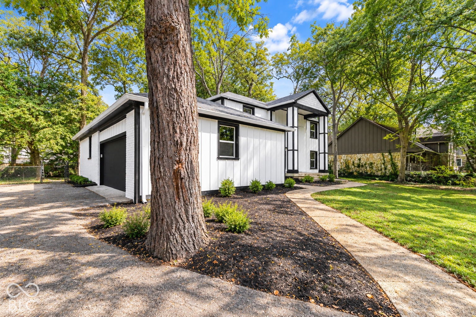 Virtually Altered - White vertical siding with black trim, integrated garage with black door, concrete walkway, mature trees, and landscaped garden beds
